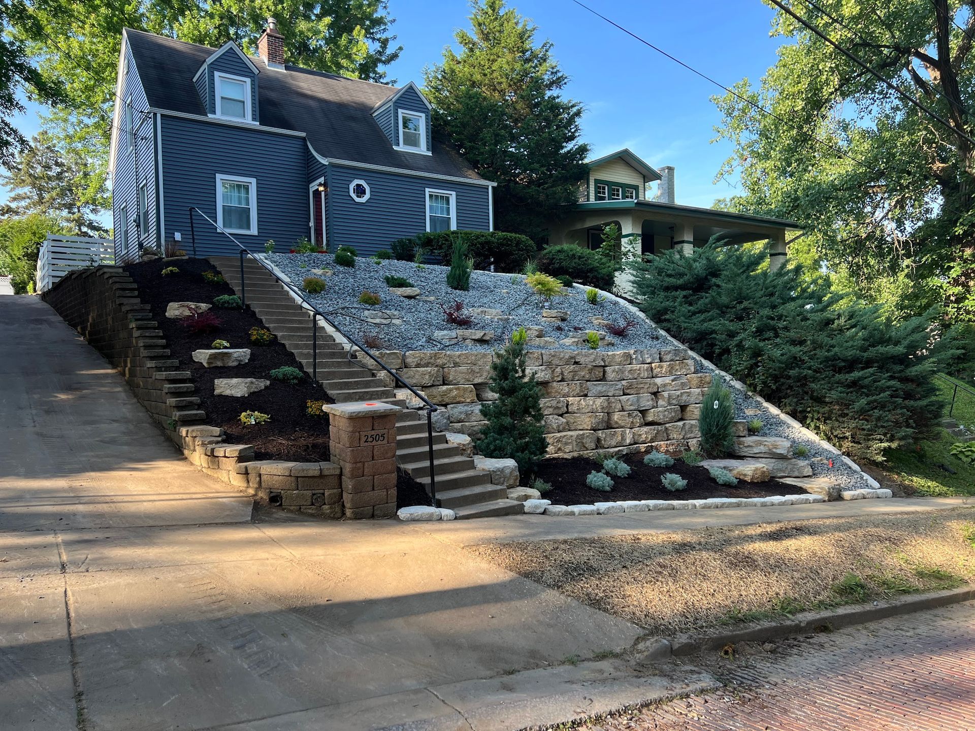 Two-story house with terraced landscaping and steps up from the street. Gray stone walls and dark mulch are visible.