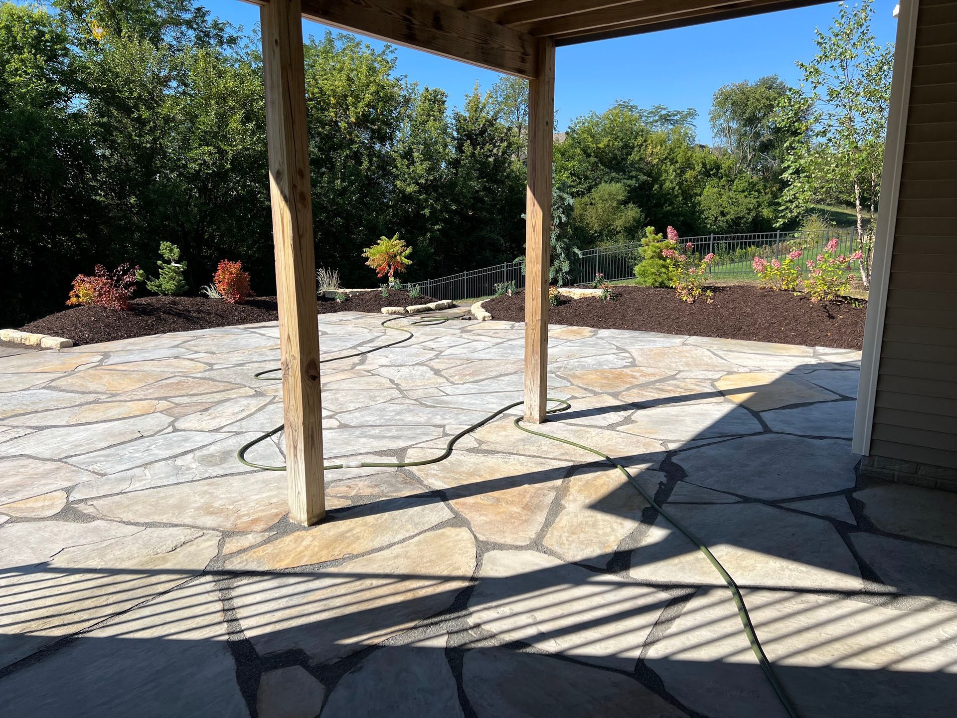 Flagstone patio with wooden supports, flower beds, and a grassy hill under a blue sky.