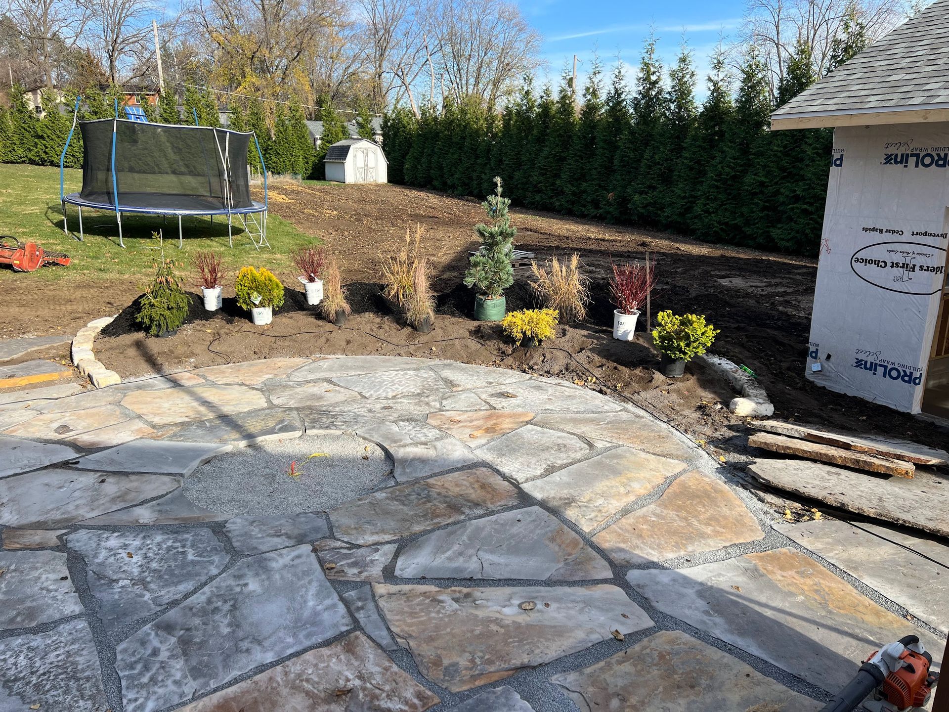 Backyard with flagstone patio and newly planted flowers, with a trampoline and hedge.