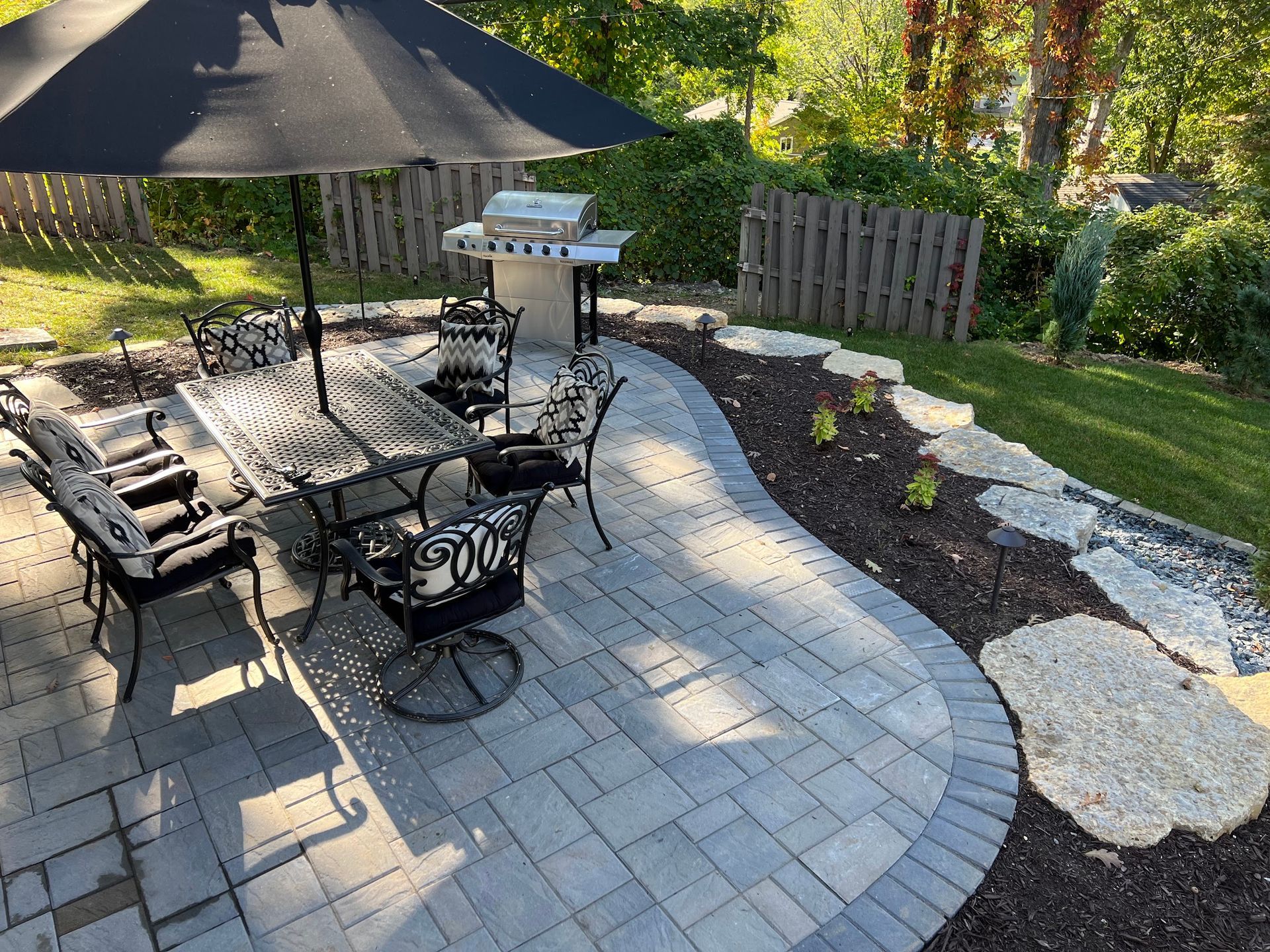 Patio with dining set, umbrella, grill, and landscaped yard. Gray pavers, black and white furniture.