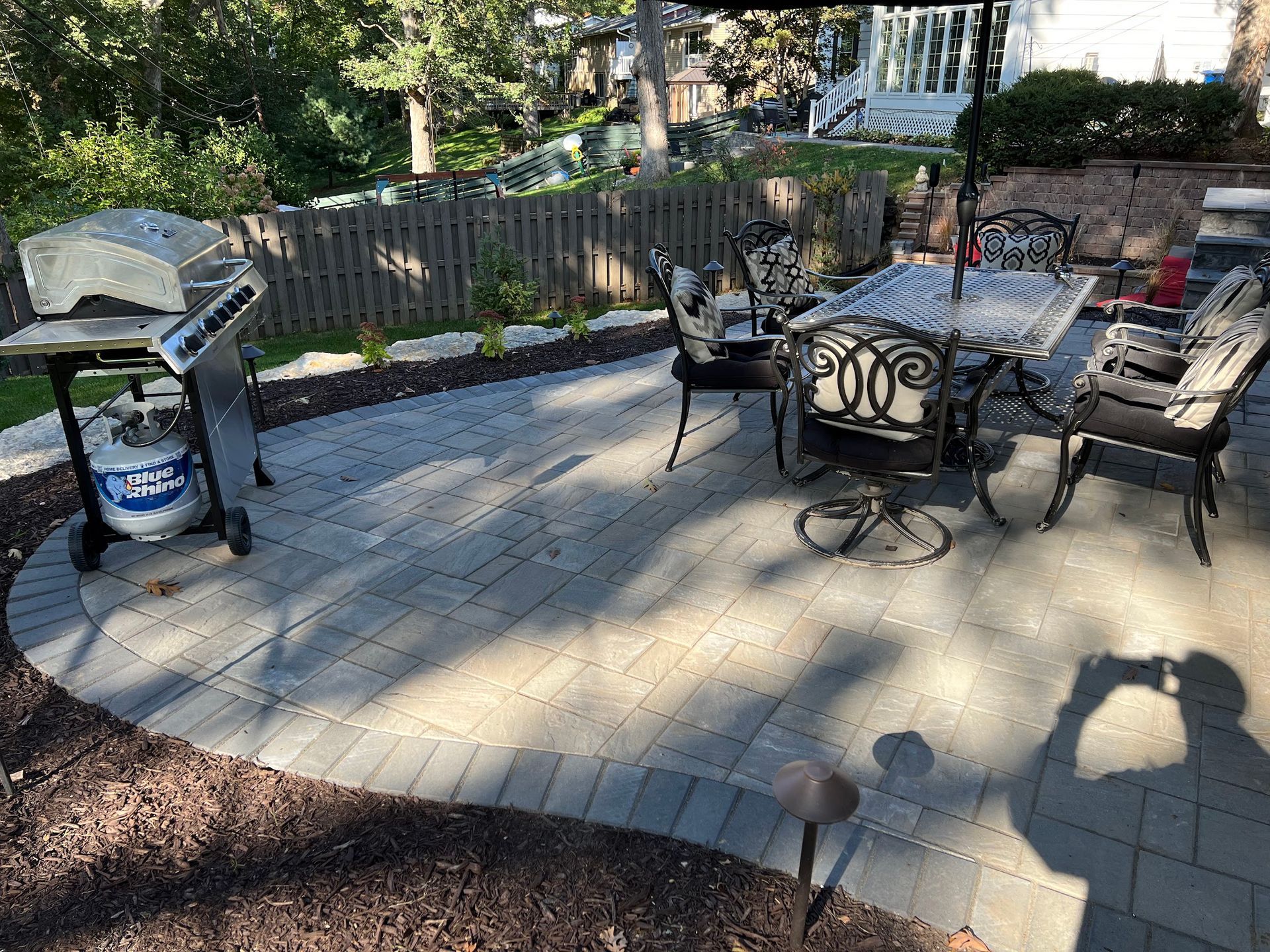 Patio with grill, table, and chairs. Paved with gray bricks, surrounded by mulch and a fence.
