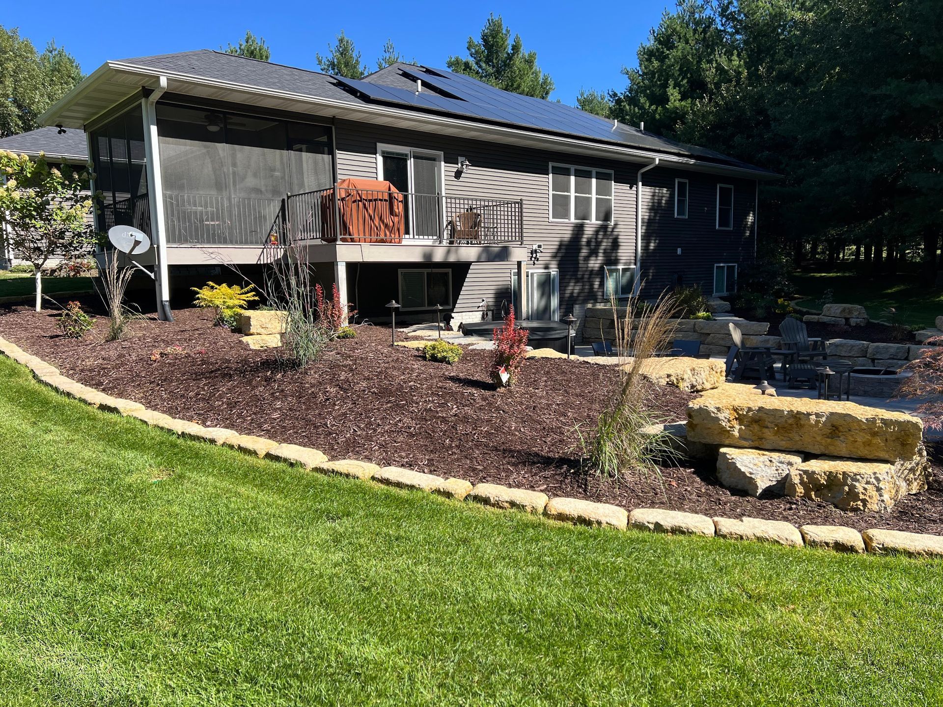 Backyard view of a house with solar panels, mulch bed, stone accents, and green grass.