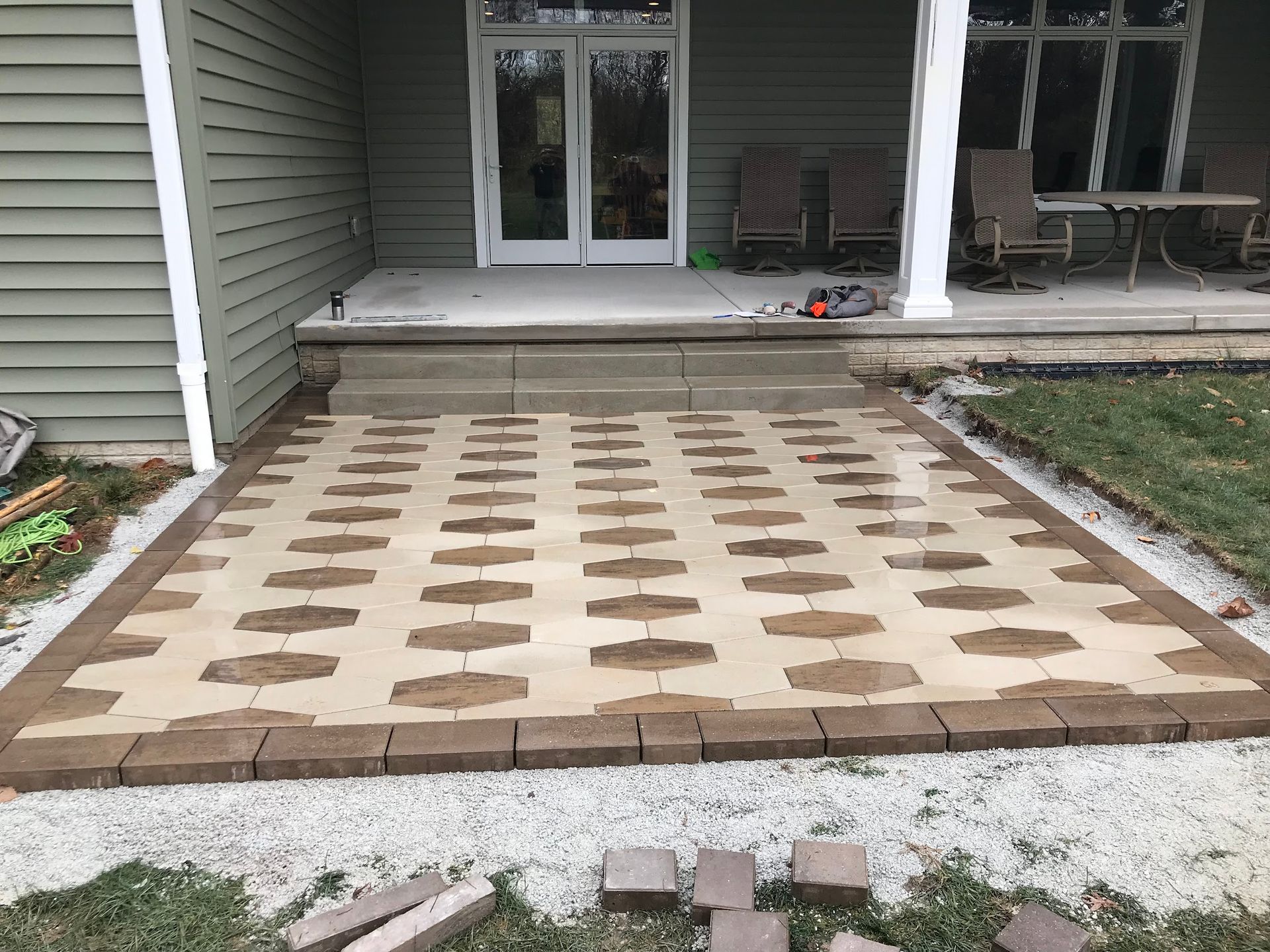 Brick patio with decorative pattern, brown border, in front of a house with steps and door.