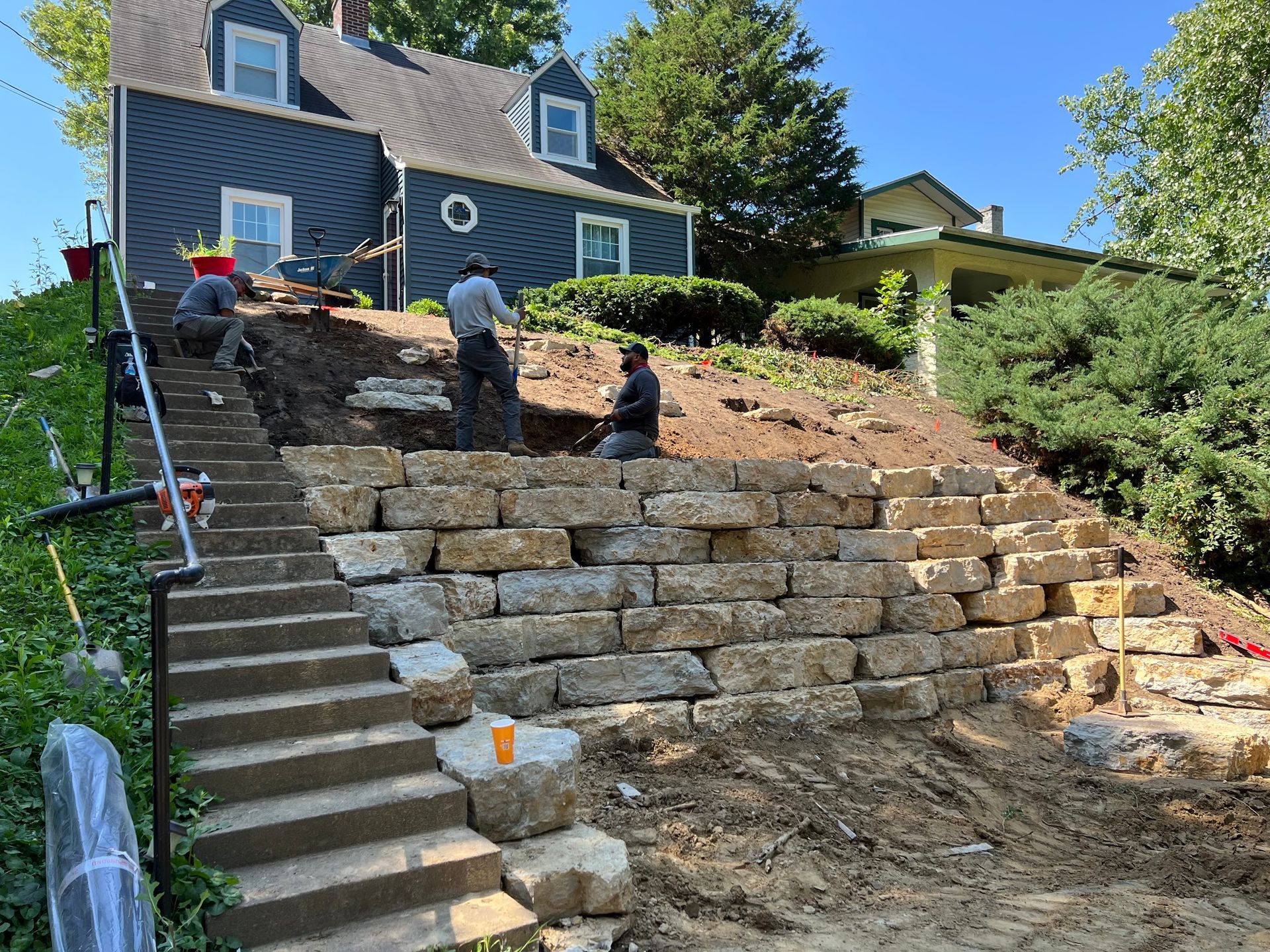 Workers building a stone retaining wall on a hillside with stairs leading up to a blue house.