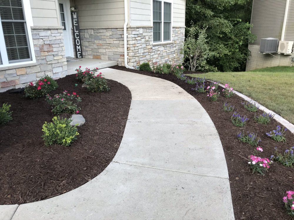 Curved concrete walkway lined with flowerbeds and mulch, leading to a house with a stone facade.