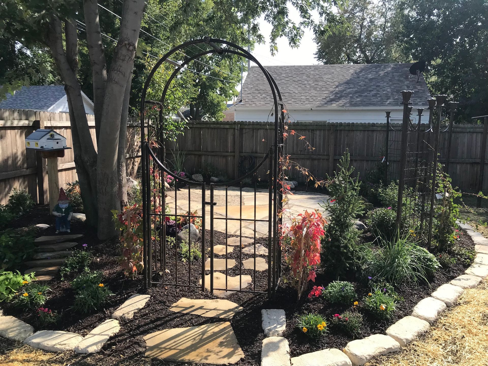 Garden with wrought-iron gate and arch, stepping stones, flowerbeds, and a wooden fence.
