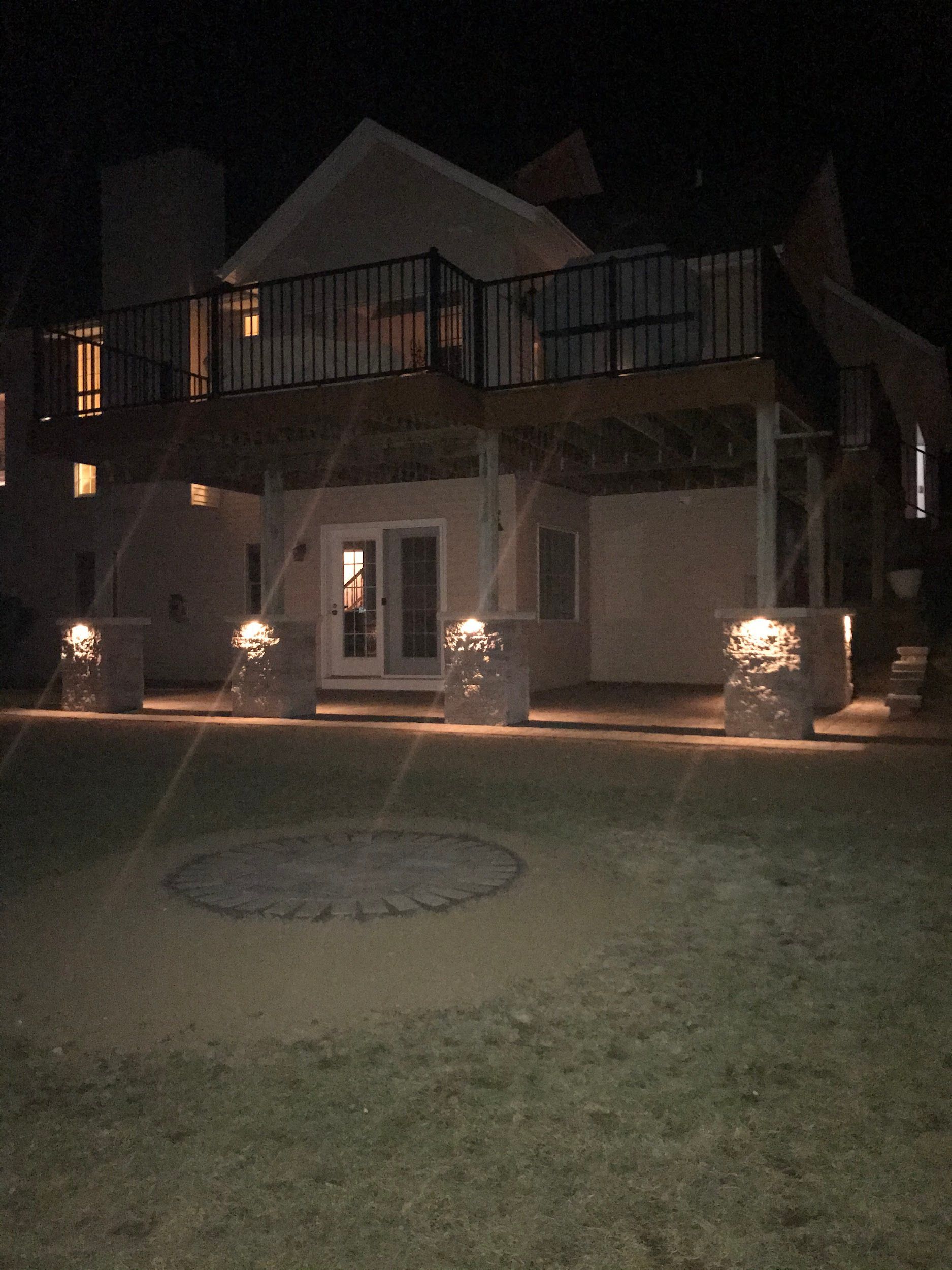 A house at night with outdoor lights illuminating pillars and a deck. A fire pit is in the foreground.