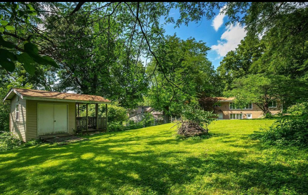 Lush green backyard with a shed, trees, and house in the distance under a partly cloudy sky.