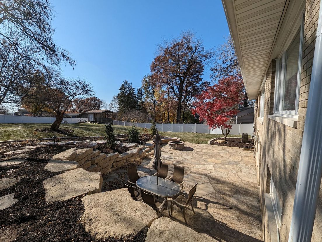 Backyard patio with stone path, dining set, and colorful autumn trees under a blue sky.