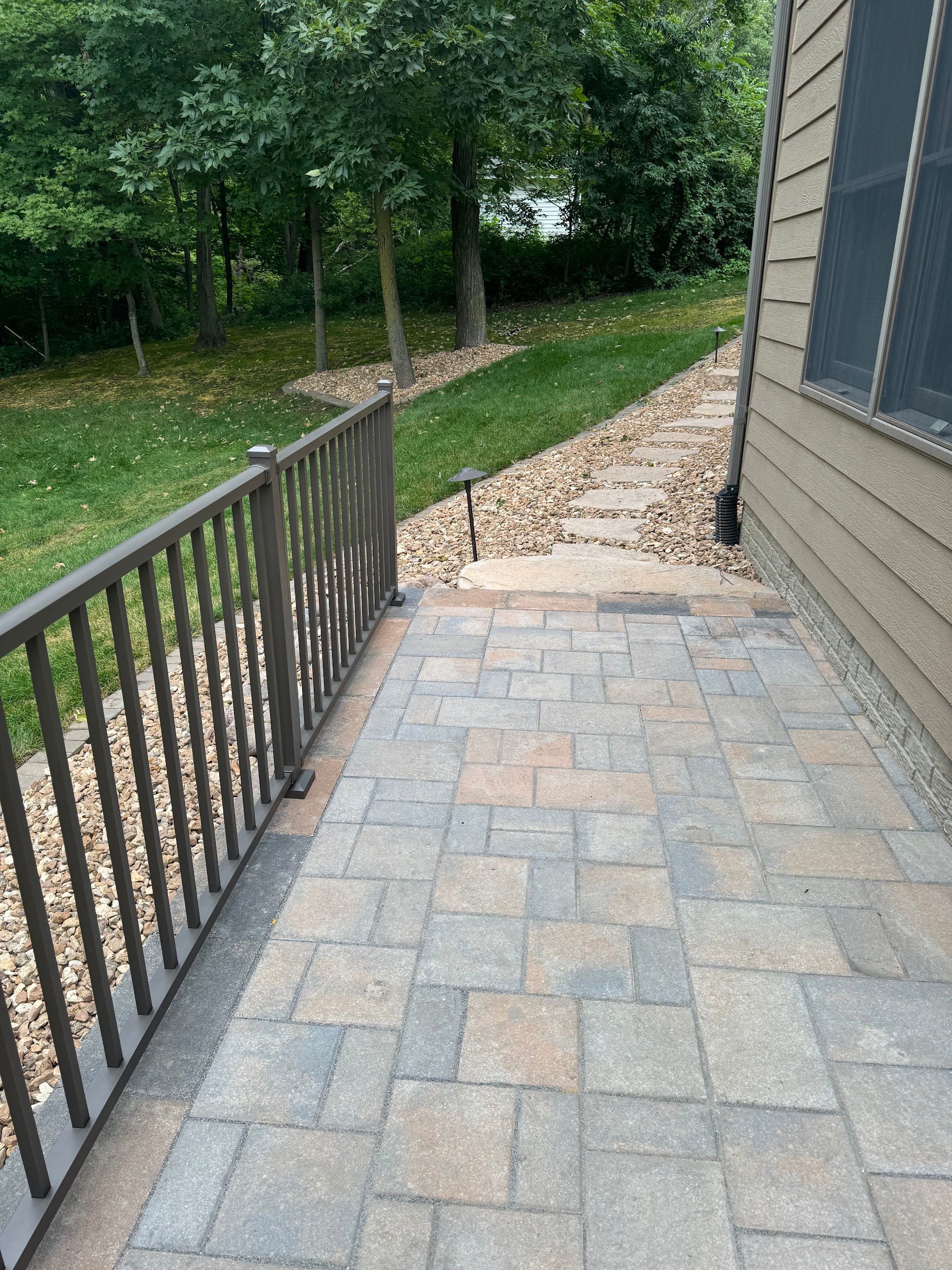 Brick patio next to a house with a metal railing and steps leading to a gravel path.