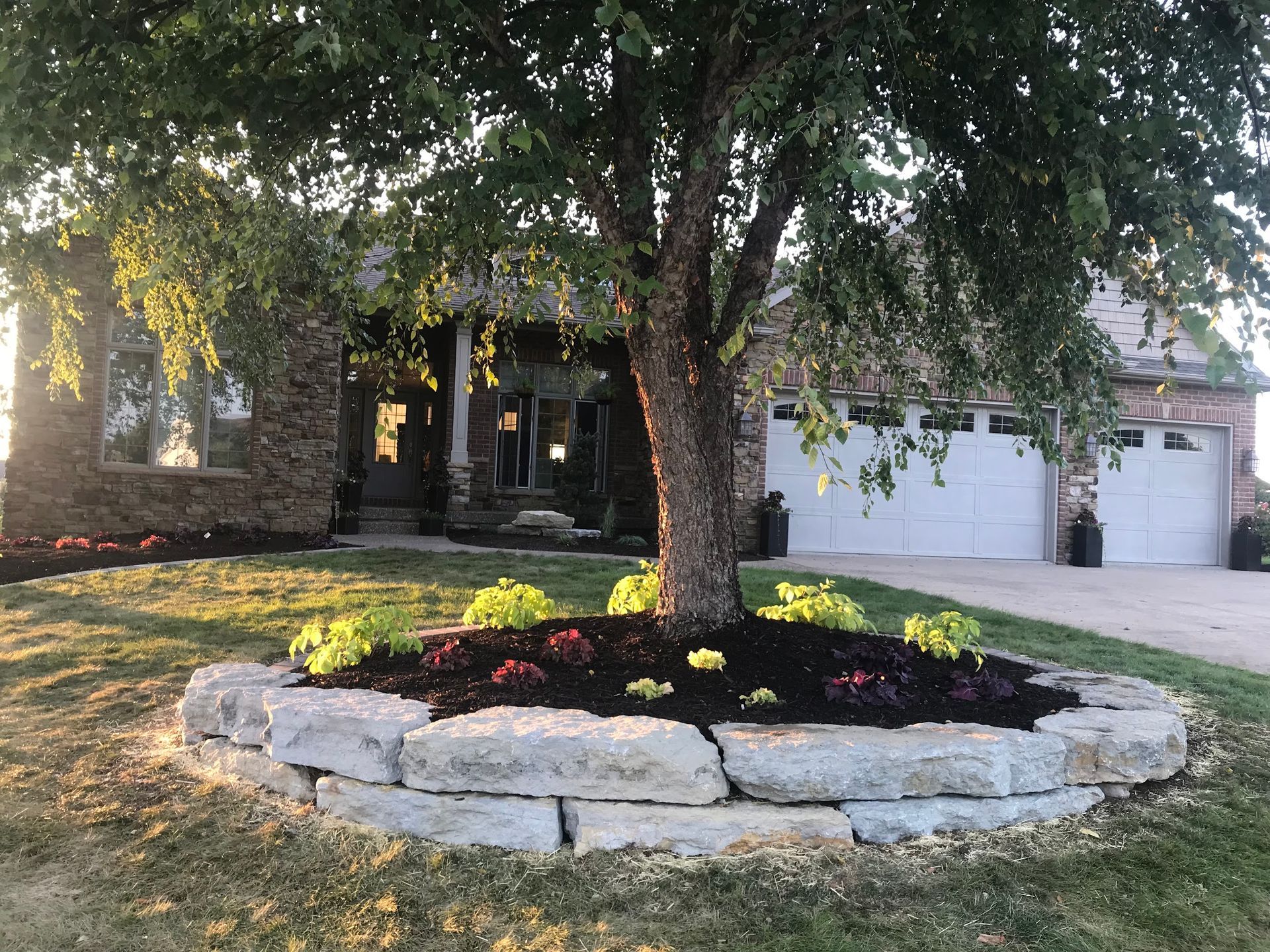 House with stone facade, tree in the front yard with a stone border, and flower bed.