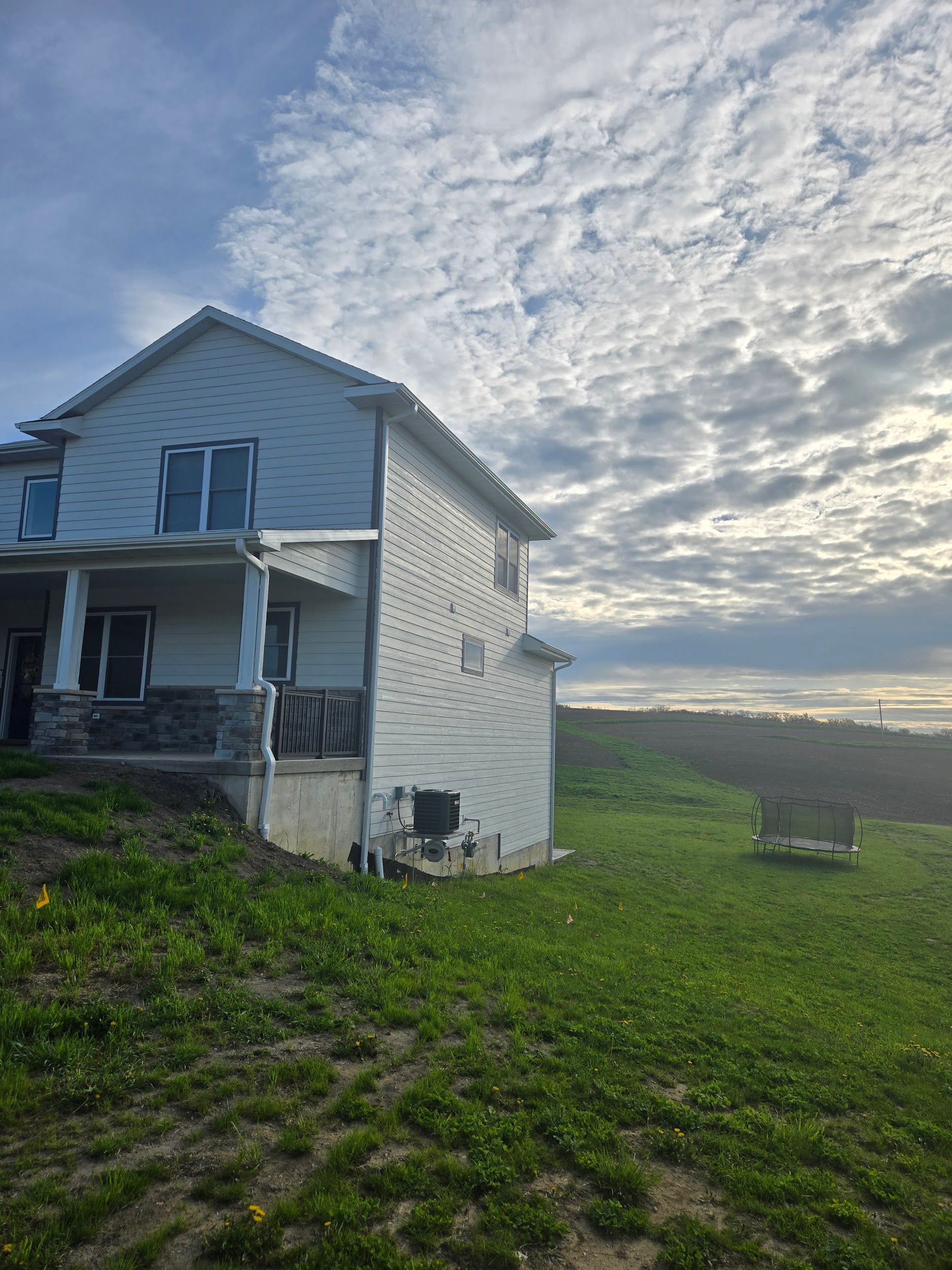 Two-story house with blue siding, porch, and grassy hill under a cloudy sky.