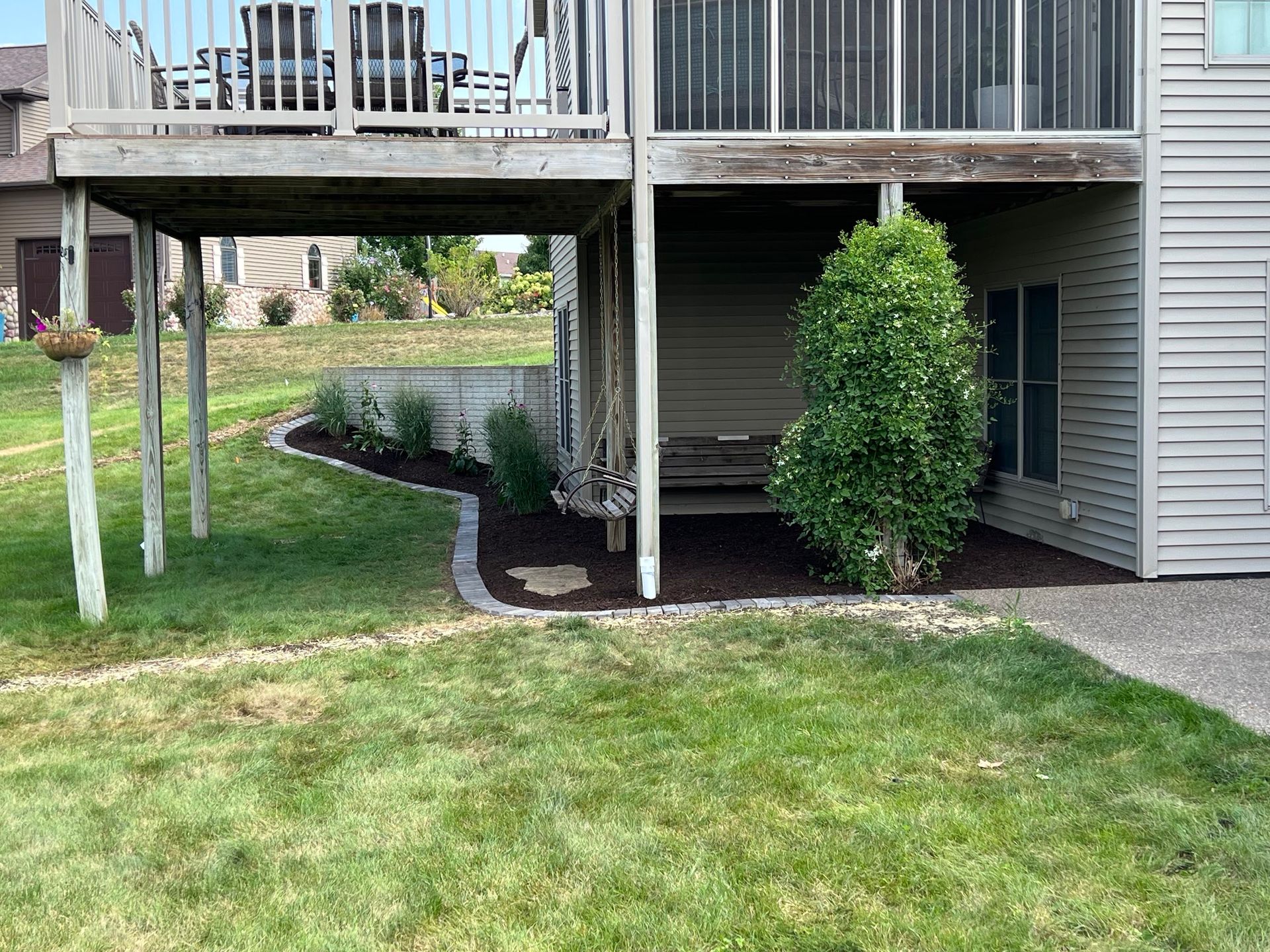 A deck overhangs a landscaped area with mulch, plants, and a tree, beside a building with sliding glass doors.