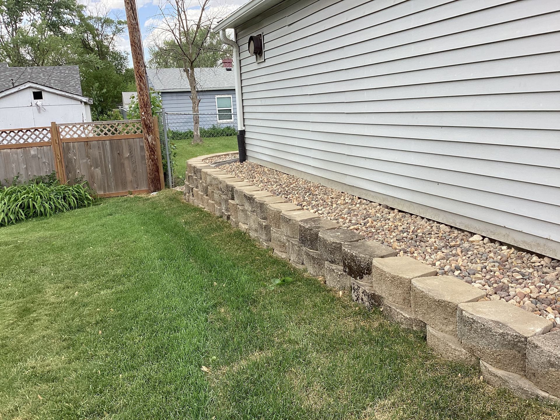Stone retaining wall next to a house with siding, gravel and grass.