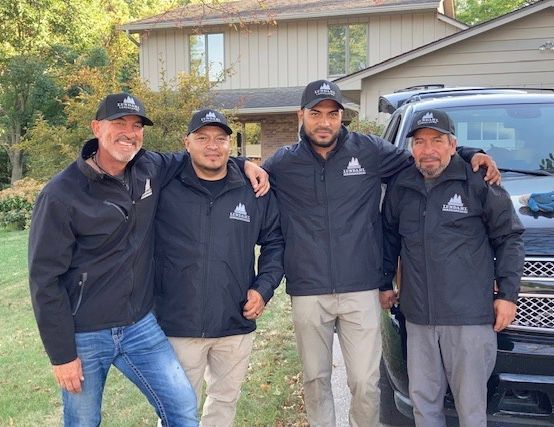 Four men in black jackets and hats pose in front of a house, smiling near a black SUV.
