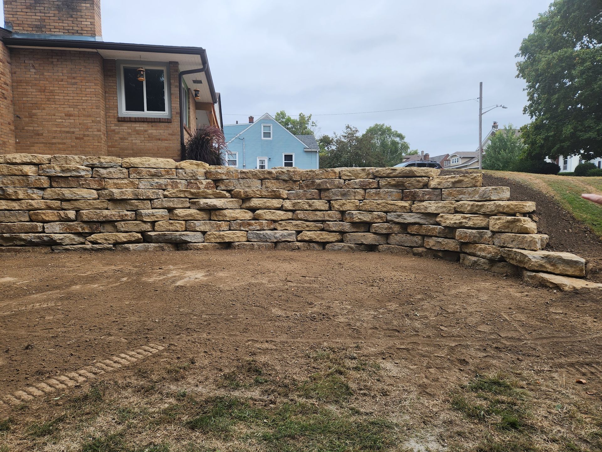 A tan stone retaining wall in front of a brick house; dirt yard, cloudy sky.
