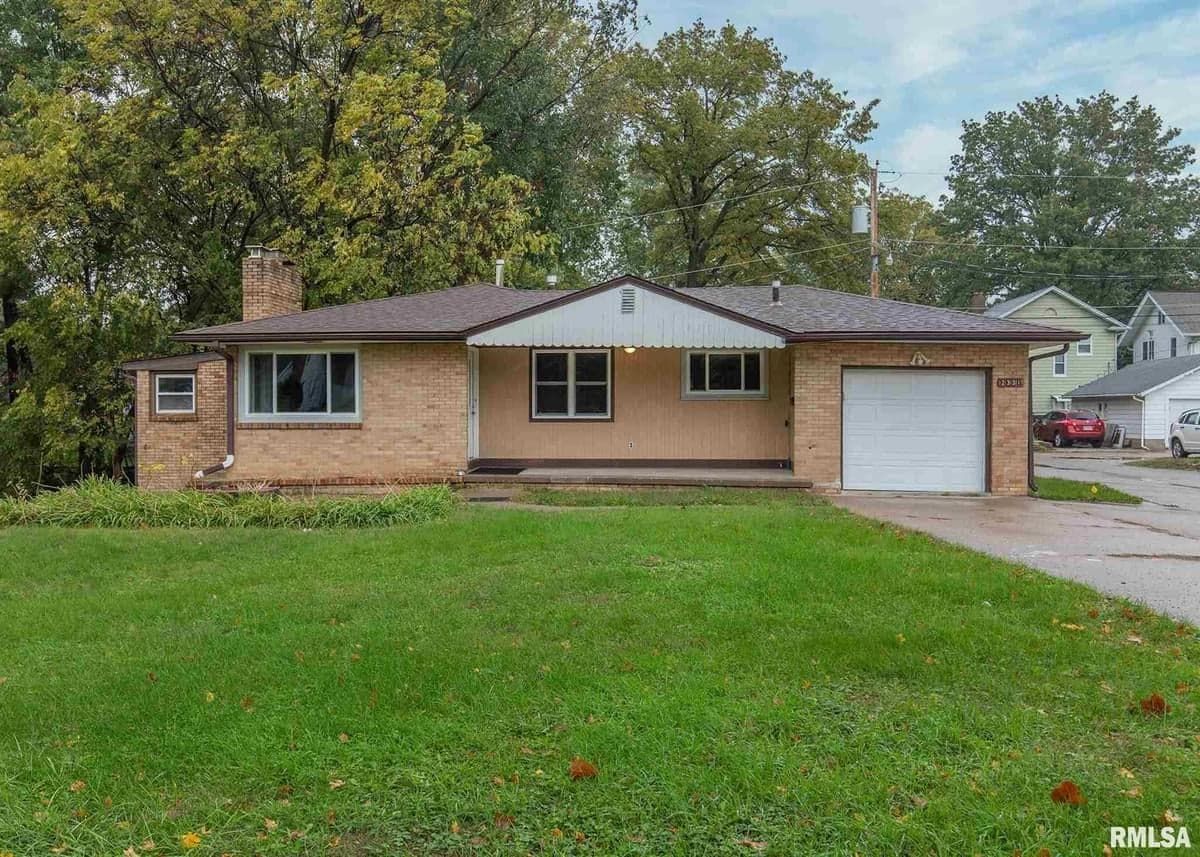 Single-story brick home with a green lawn, garage, and trees in the background under a cloudy sky.