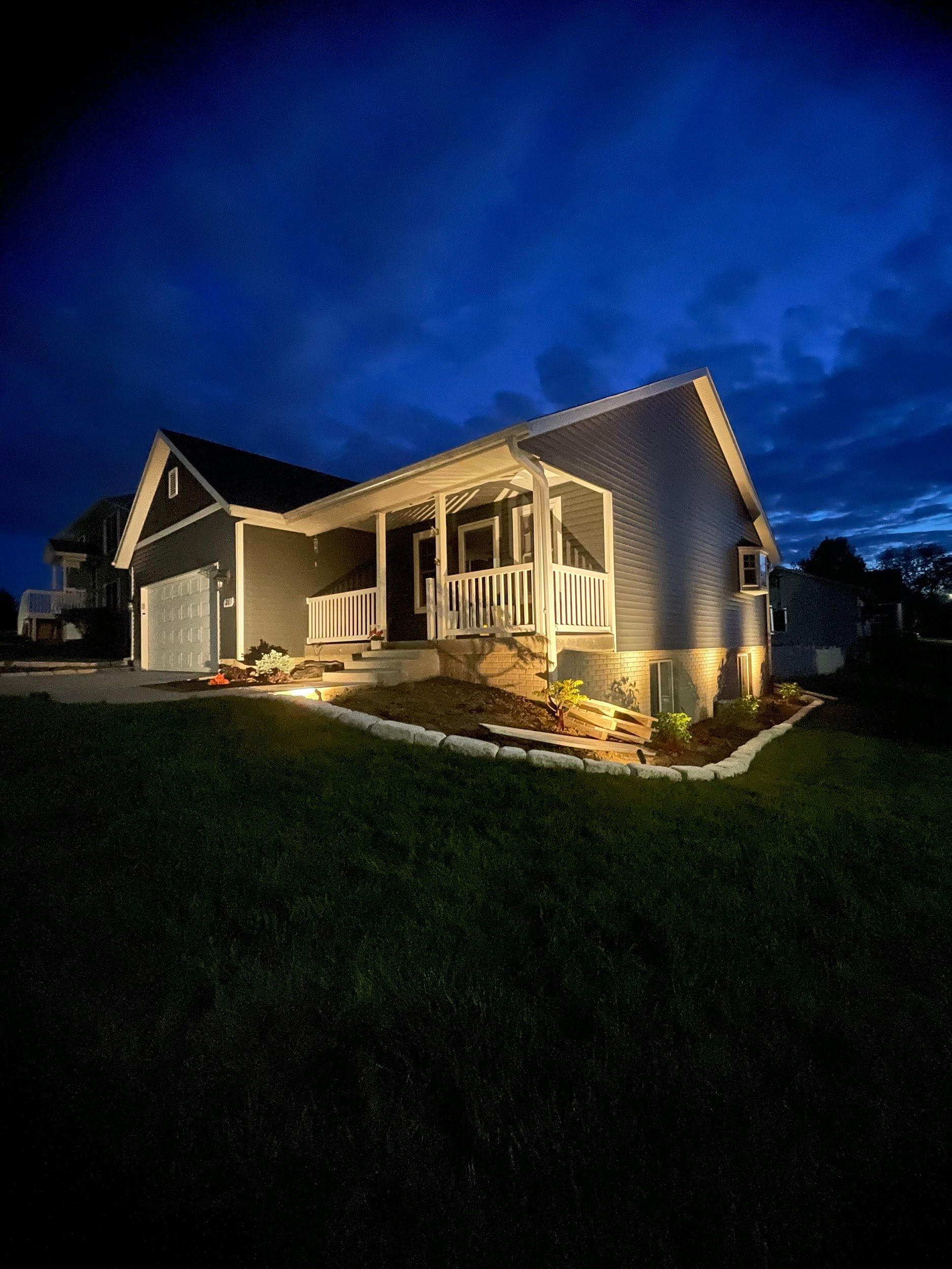 House at dusk with porch lights, dark blue siding, and green lawn.