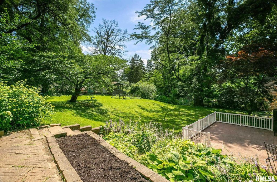 Lush green backyard with stairs, a garden bed, and trees under a sunny sky.