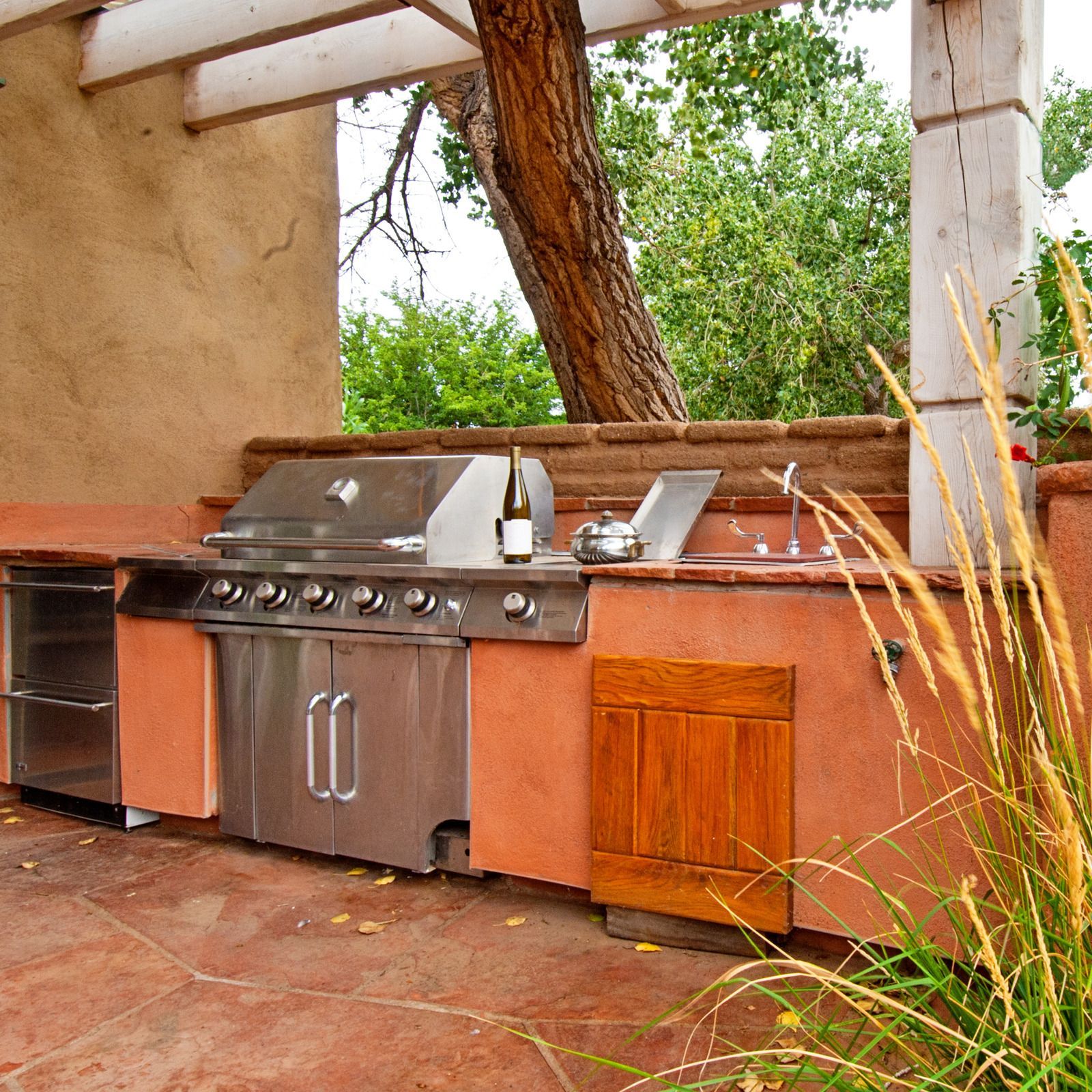 Outdoor kitchen with stainless steel grill, sink, and refrigerator; built-in, with red-orange stucco, near a tree.