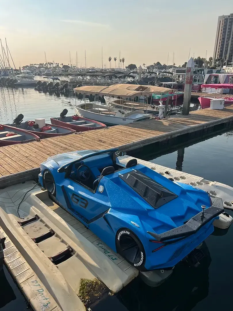 A blue sports car is parked on a dock next to a body of water.