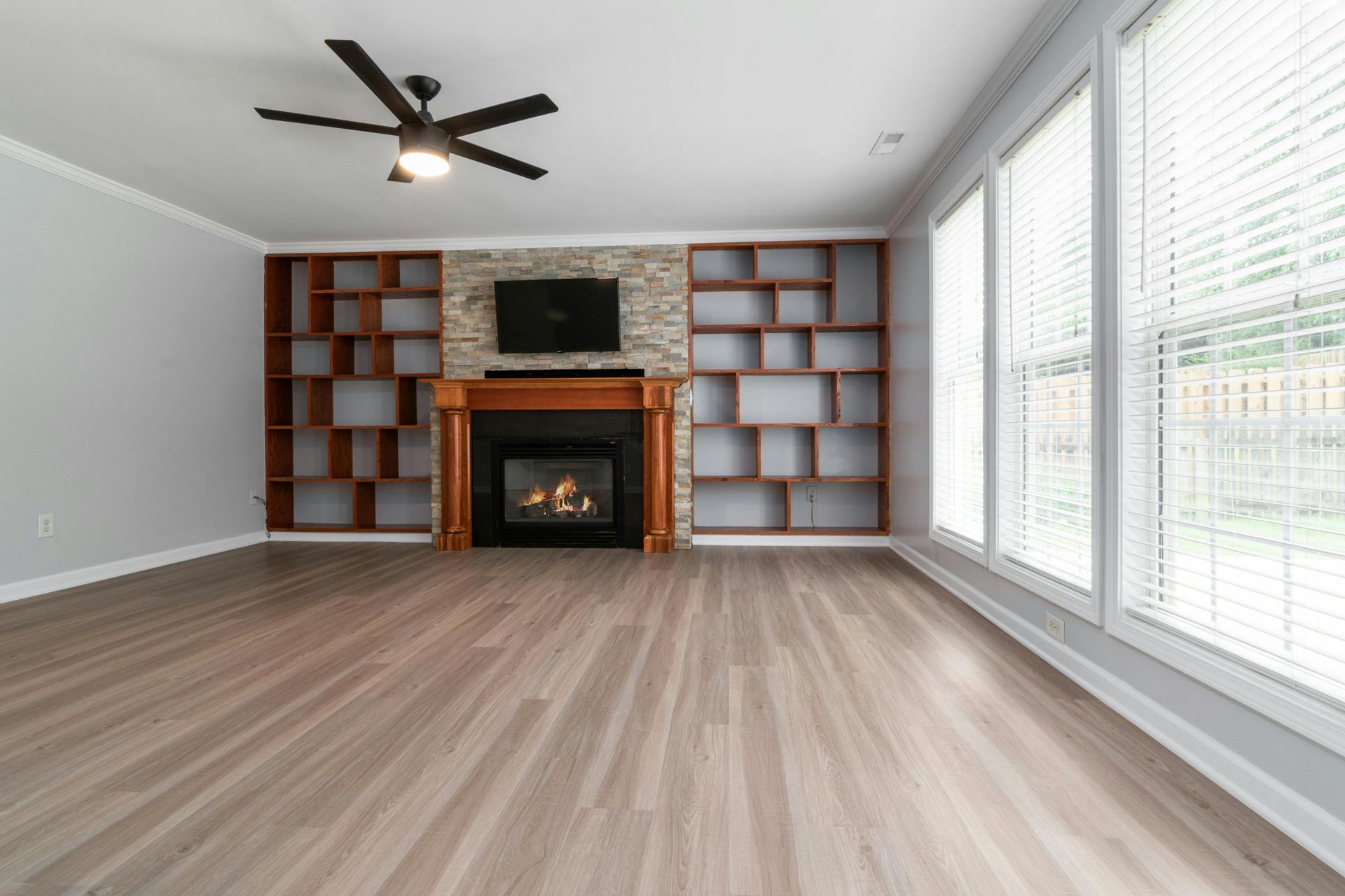 Empty living room with fireplace, built-in shelves, ceiling fan, and large windows with blinds.