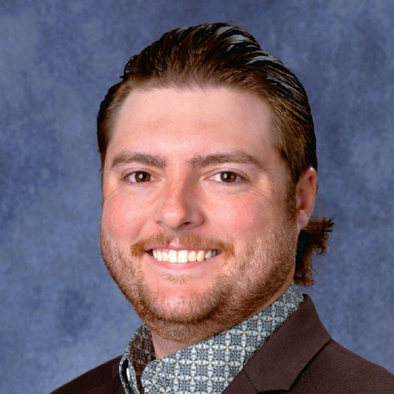 A smiling person with short, light brown hair and a beard, wearing a patterned collared shirt and blazer against a blue backdrop.
