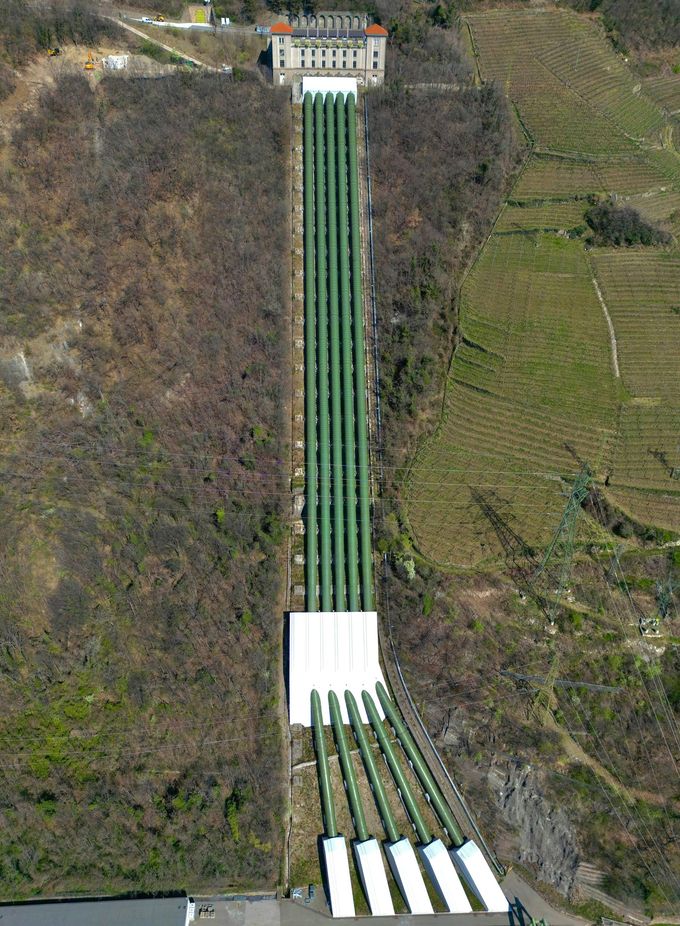 Hydroelectric plant with tall pipes running down a hillside to a white building.