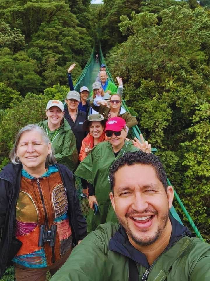 Group of hikers on a narrow suspension bridge in a lush green forest, smiling for a selfie.