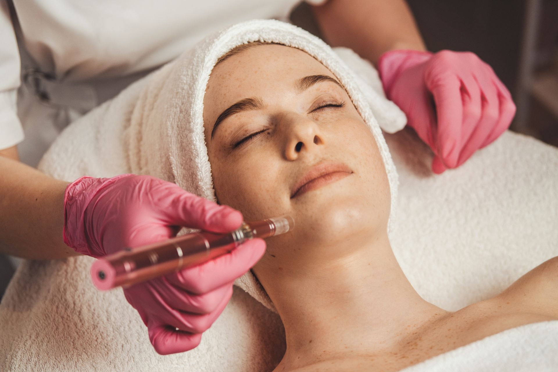 Woman receiving microneedling treatment on her face; hands in pink gloves.