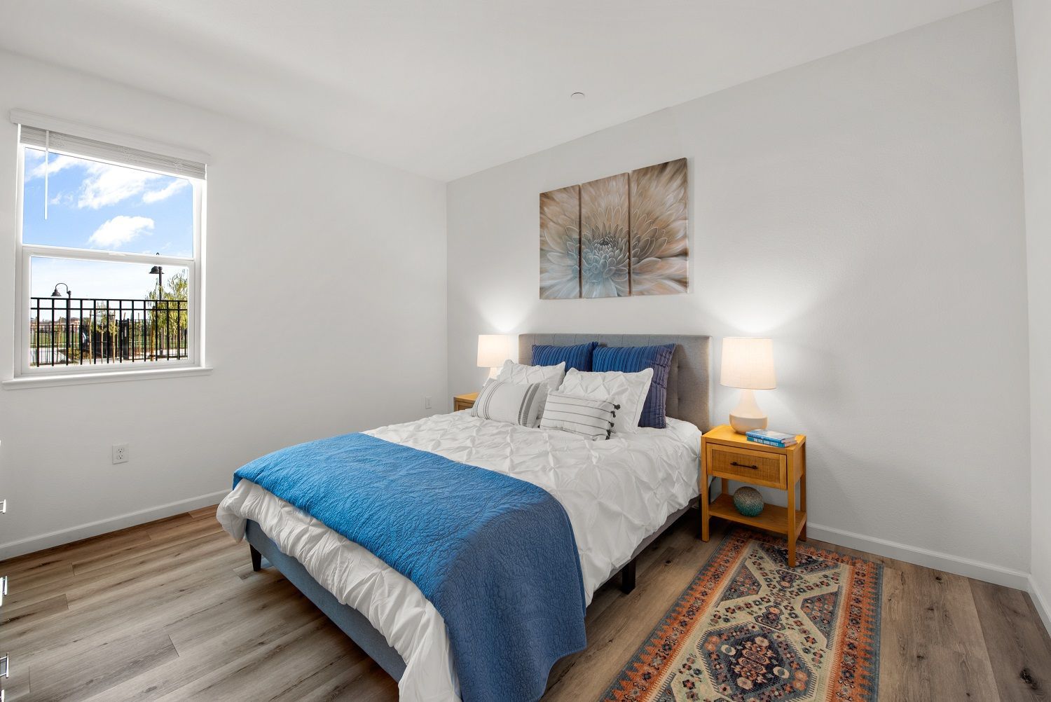 Bedroom with a queen bed, blue bedding, two nightstands, lamps, and a window. Explore floor plans designed for easy living.