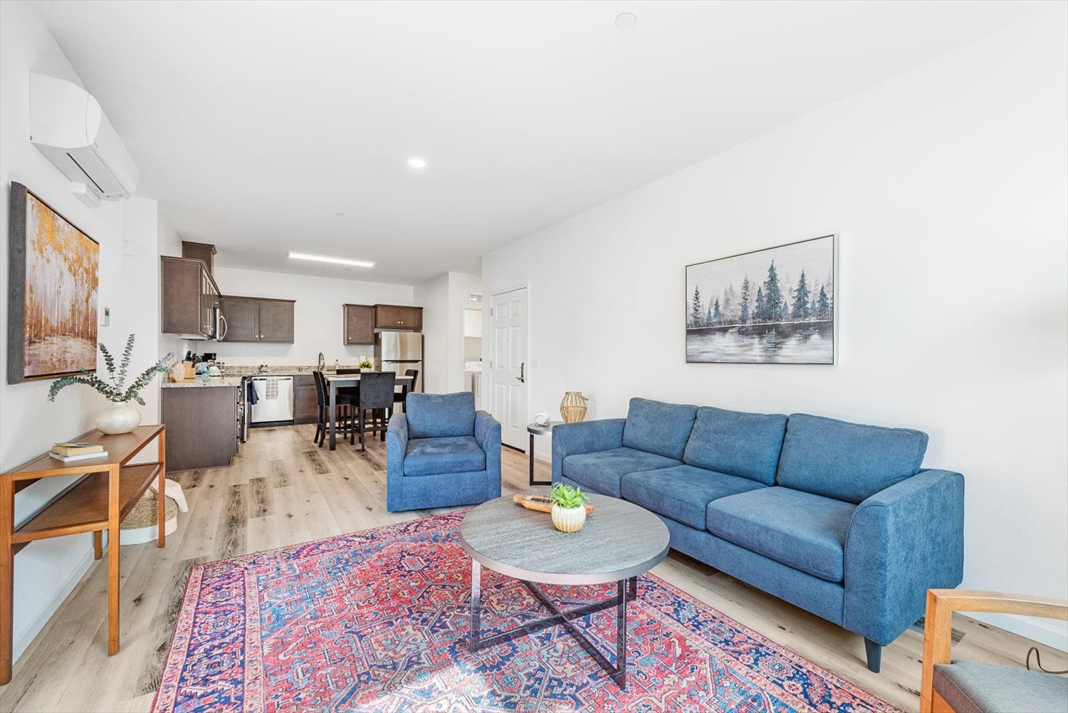 Open-concept living area with a blue sofa, matching chair, coffee table, and kitchen in background. Discover comfort in the heart of Brentwood.