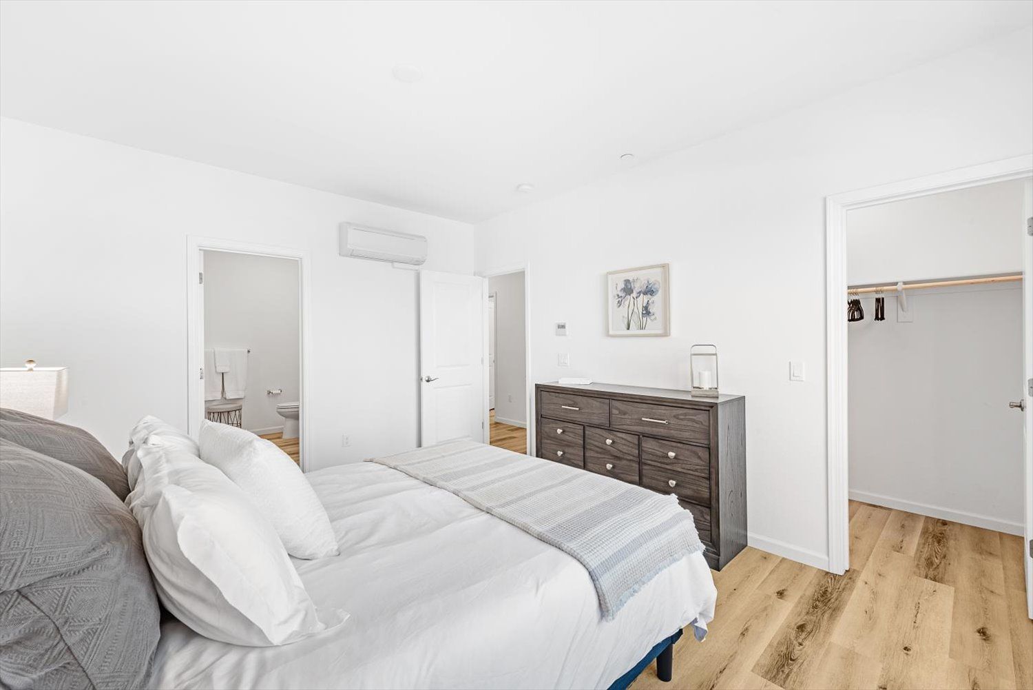 Bright bedroom with white walls, wood flooring, a large bed, dresser, and open closet. See available floor plans in Brentwood.