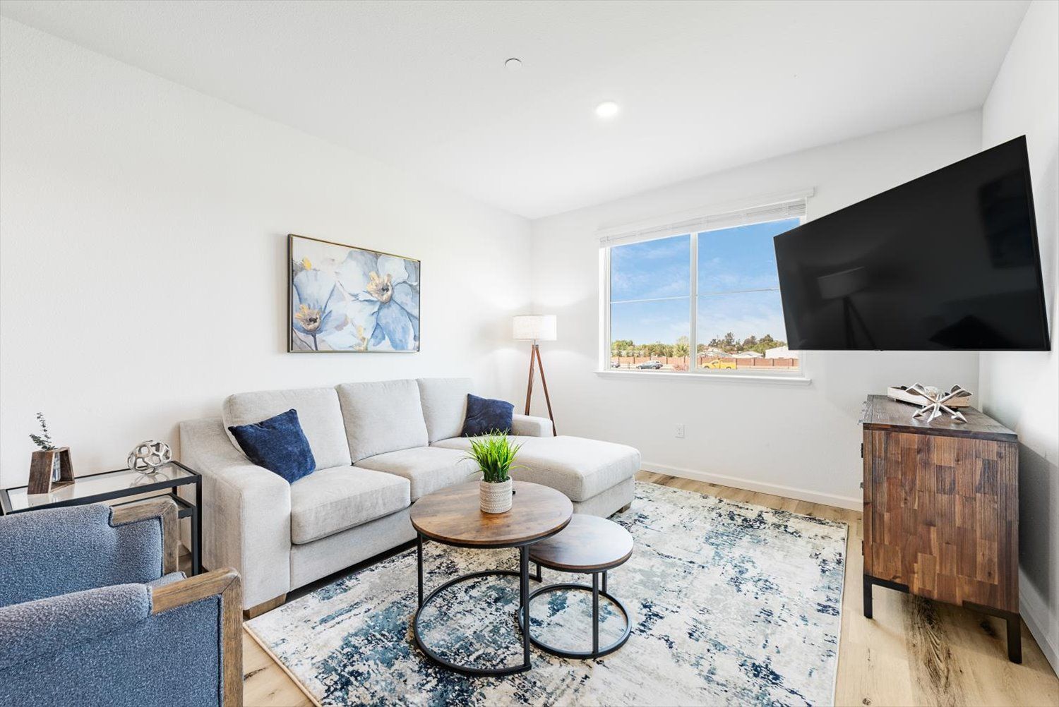 Living room in a modern apartment with a light sofa, coffee tables, rug, and a wall-mounted TV by a window. Tour The Silvergate from wherever you are.