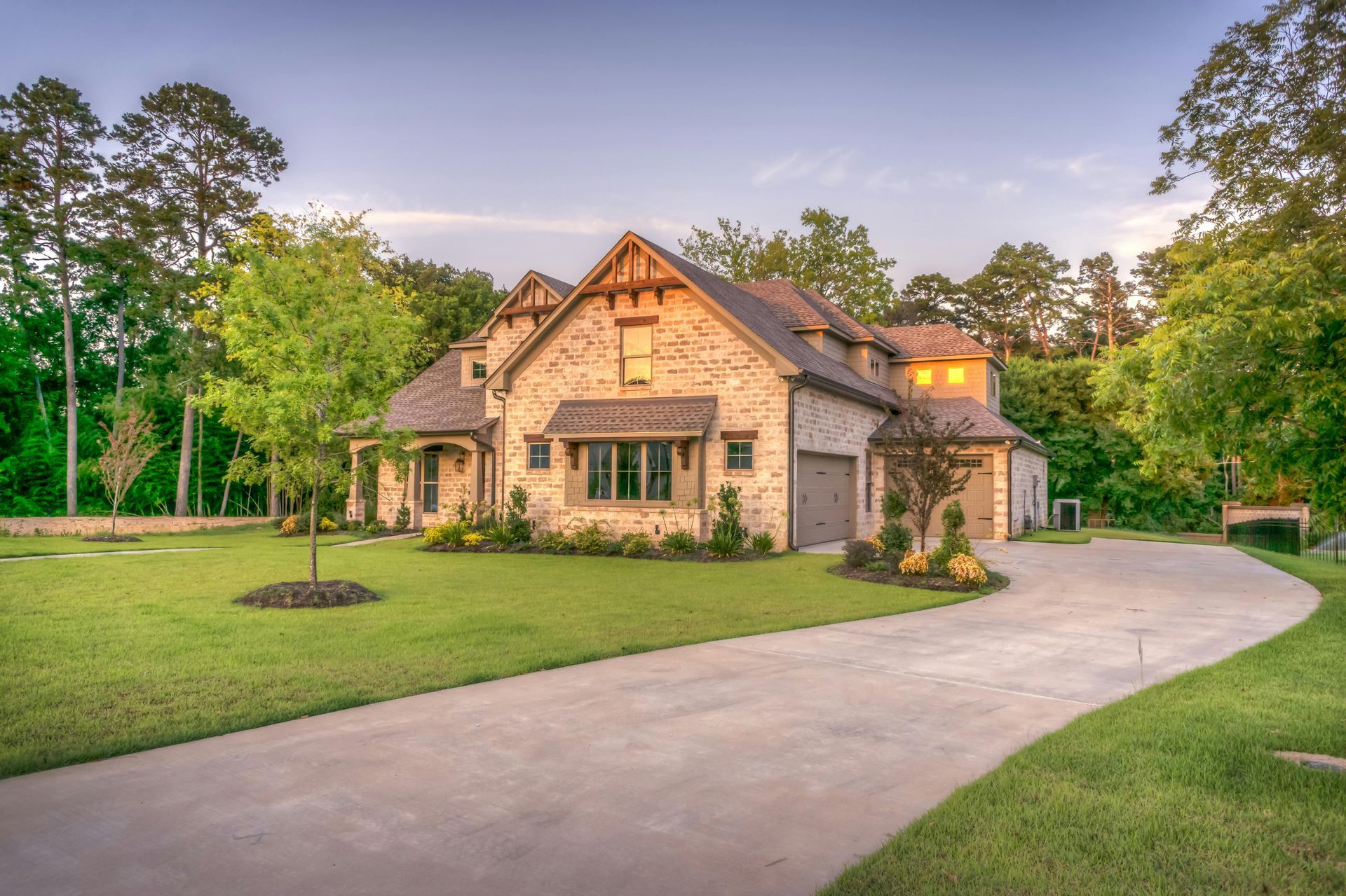 Stone house with a curved driveway, green lawn, and trees at dusk