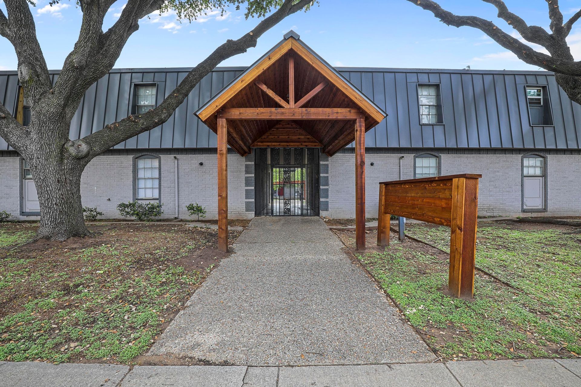 Apartment building entrance with wooden awning, walkway, and sign. Brick exterior, trees frame the view.