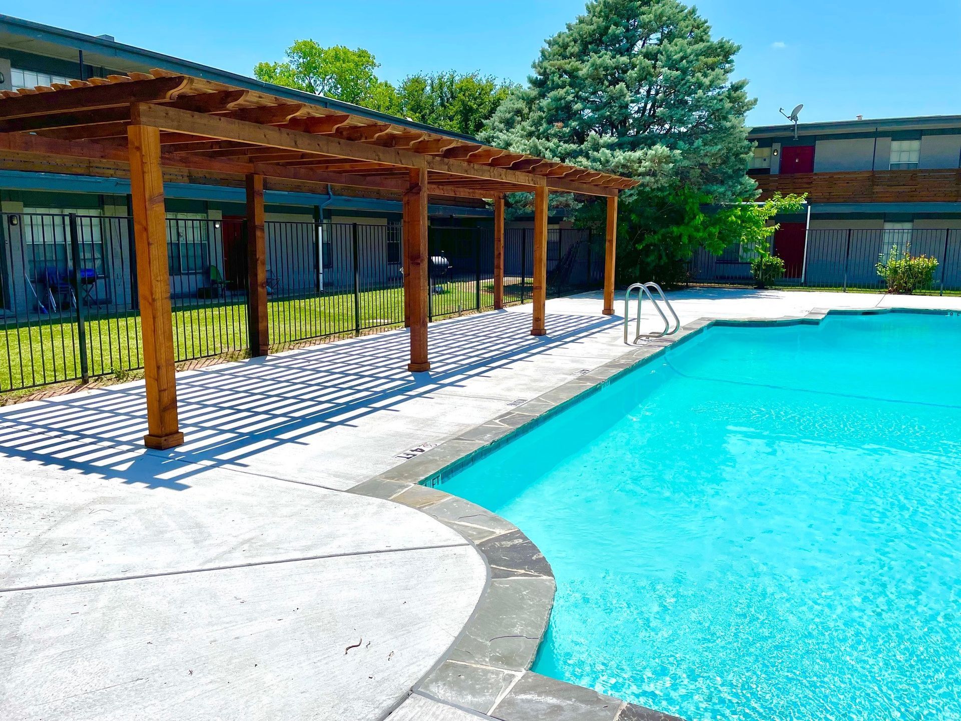 Swimming pool next to a wooden pergola with apartment building in background under a blue sky.