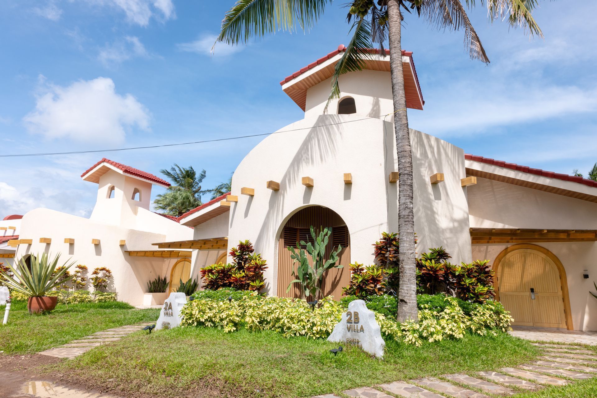 White stucco house with red tile roof, arched entry, and tropical landscaping under a blue sky