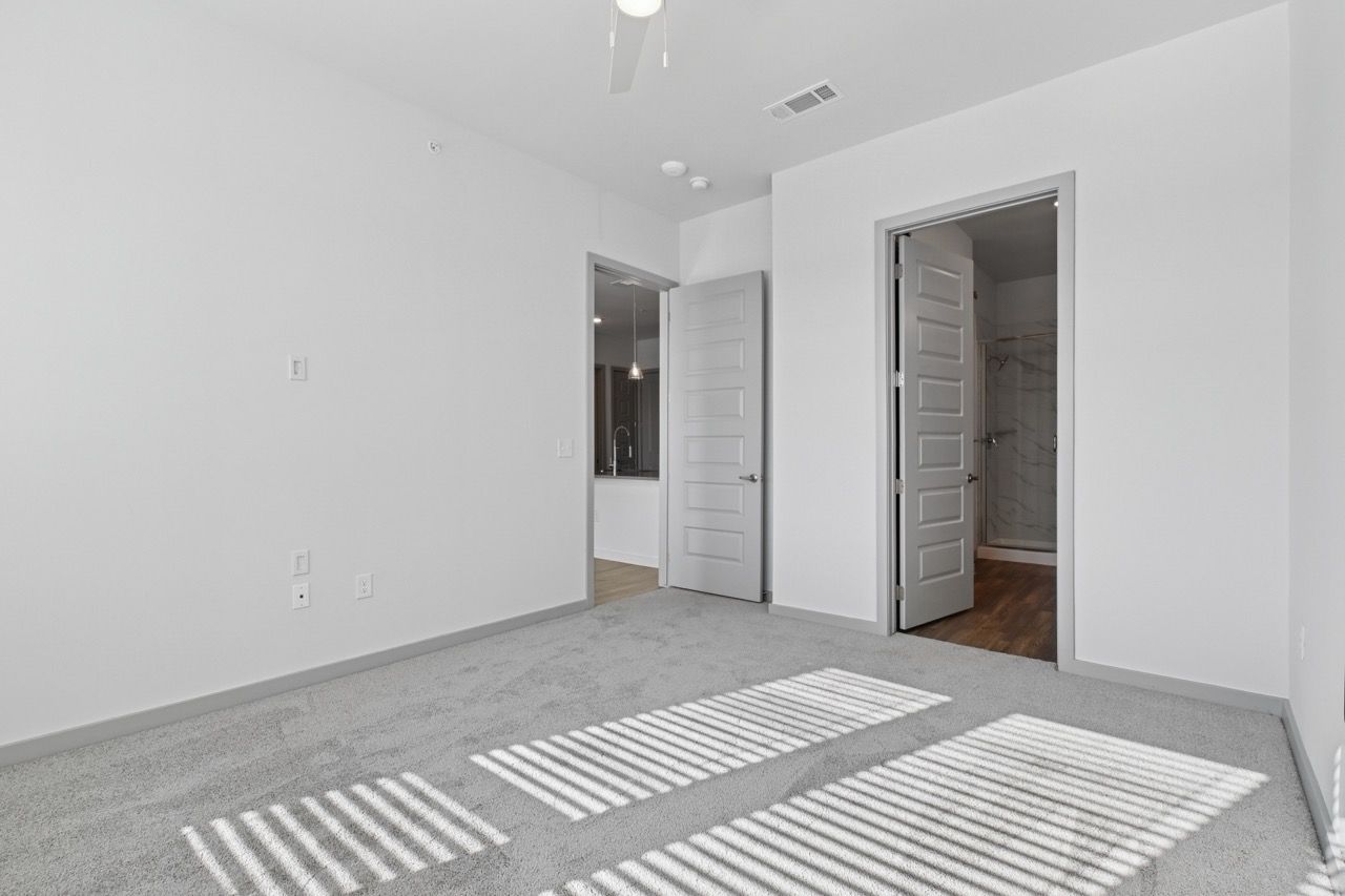 Empty bedroom with white walls, gray carpet, and two doors to a bathroom and hallway.