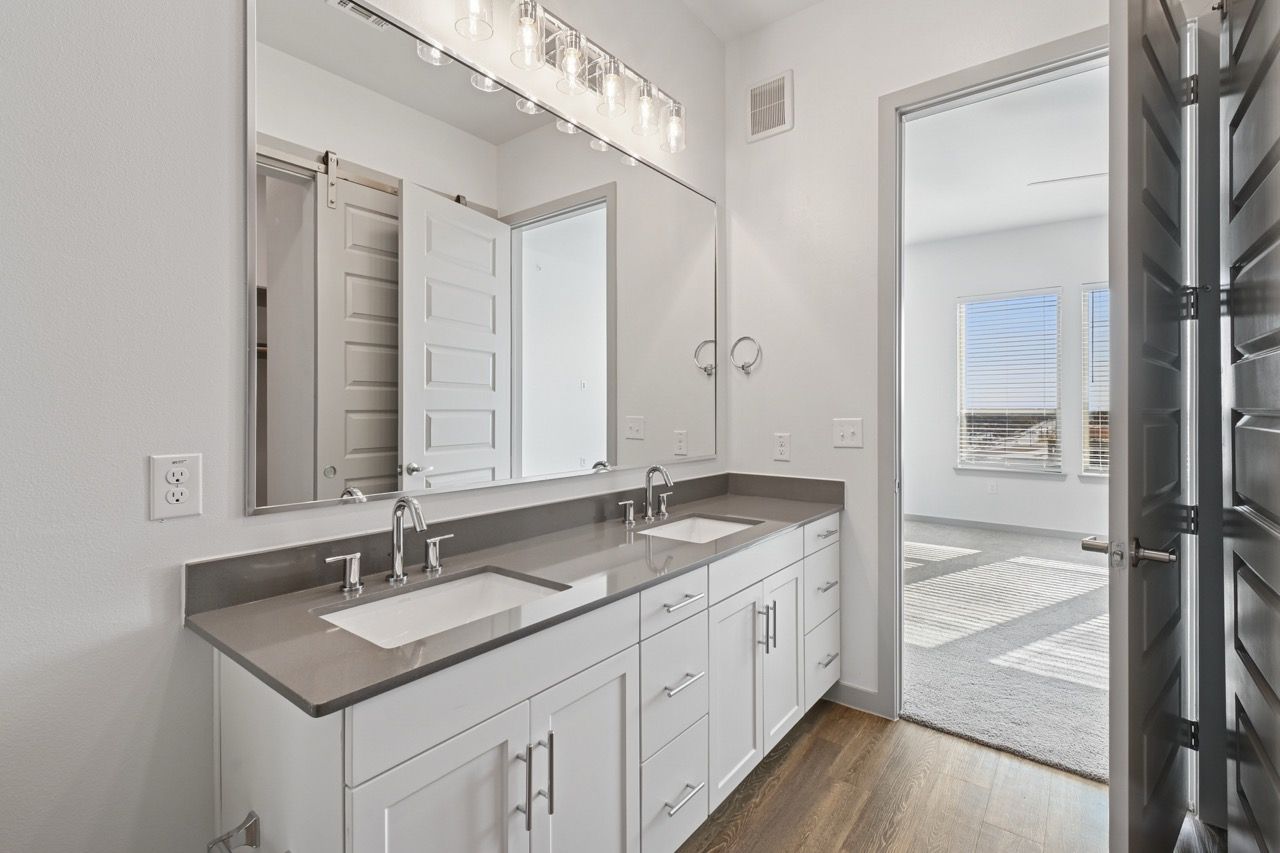 Two-sink bathroom vanity with gray countertop, large mirror, and doorway to a sunlit bedroom.