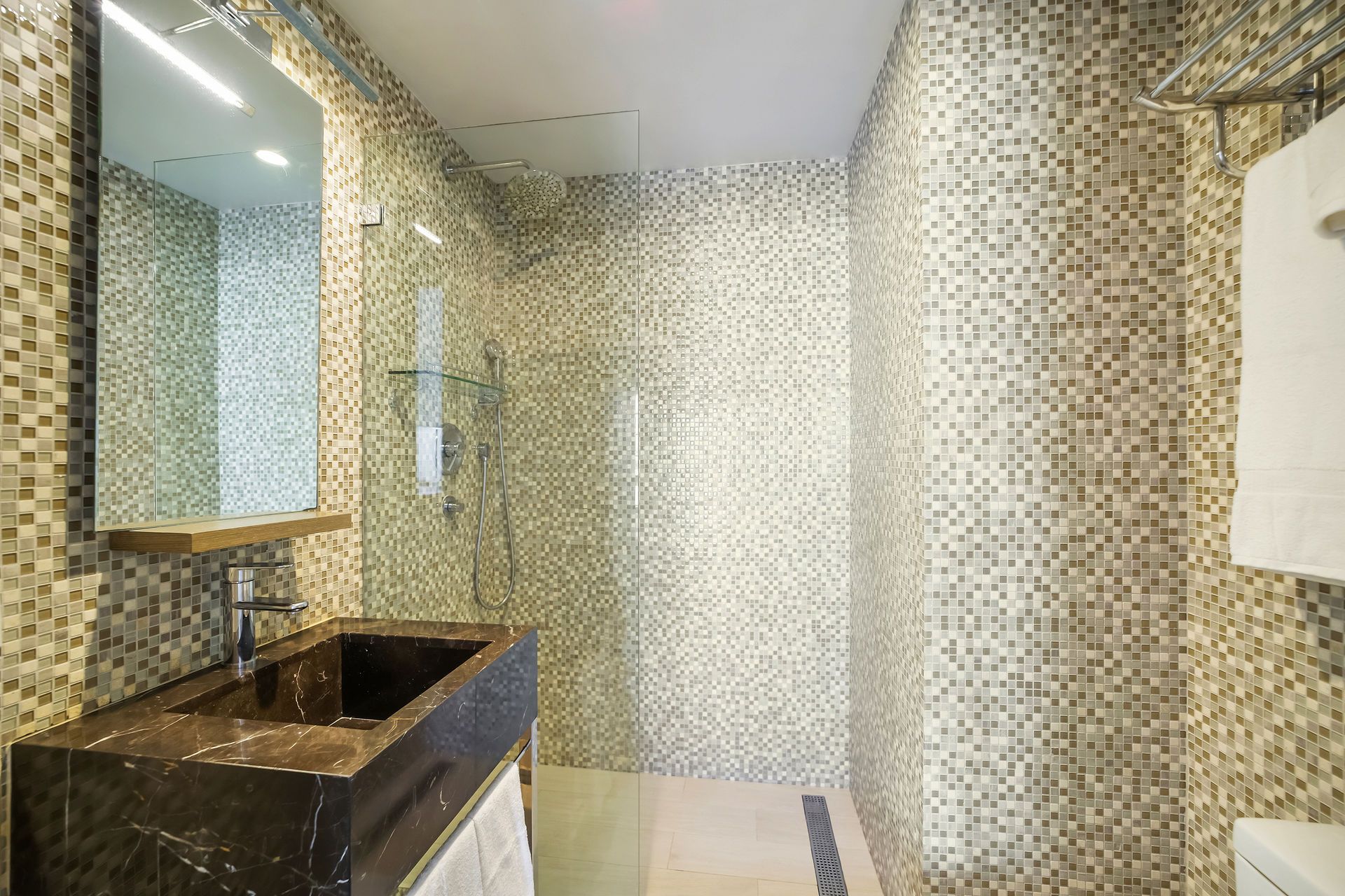 Bathroom with mosaic tiled walls, dark countertop sink, shower, and a towel on a rack.