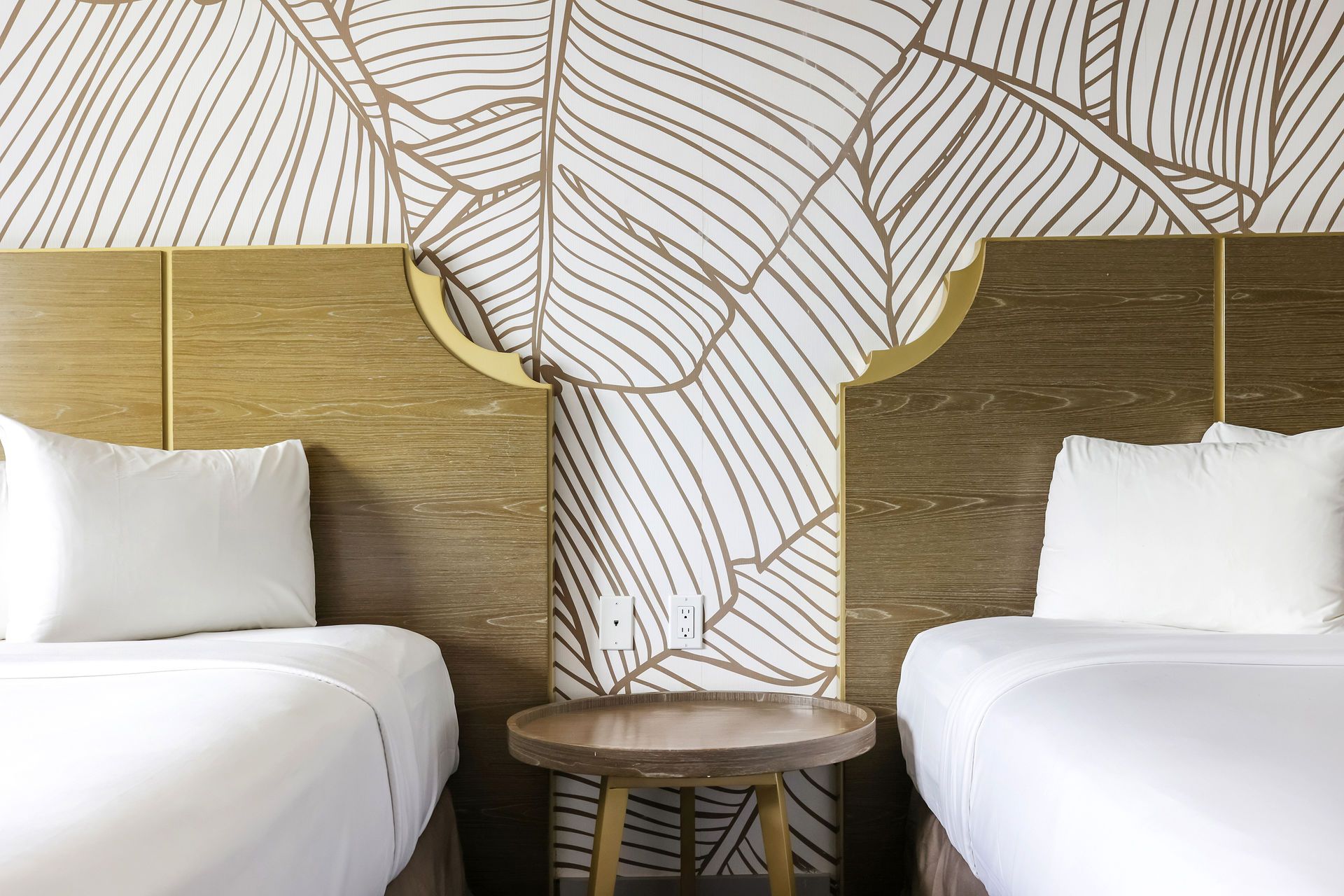 Two beds in a hotel room, with white linens and a wood-toned headboard. A small round table sits between the beds.