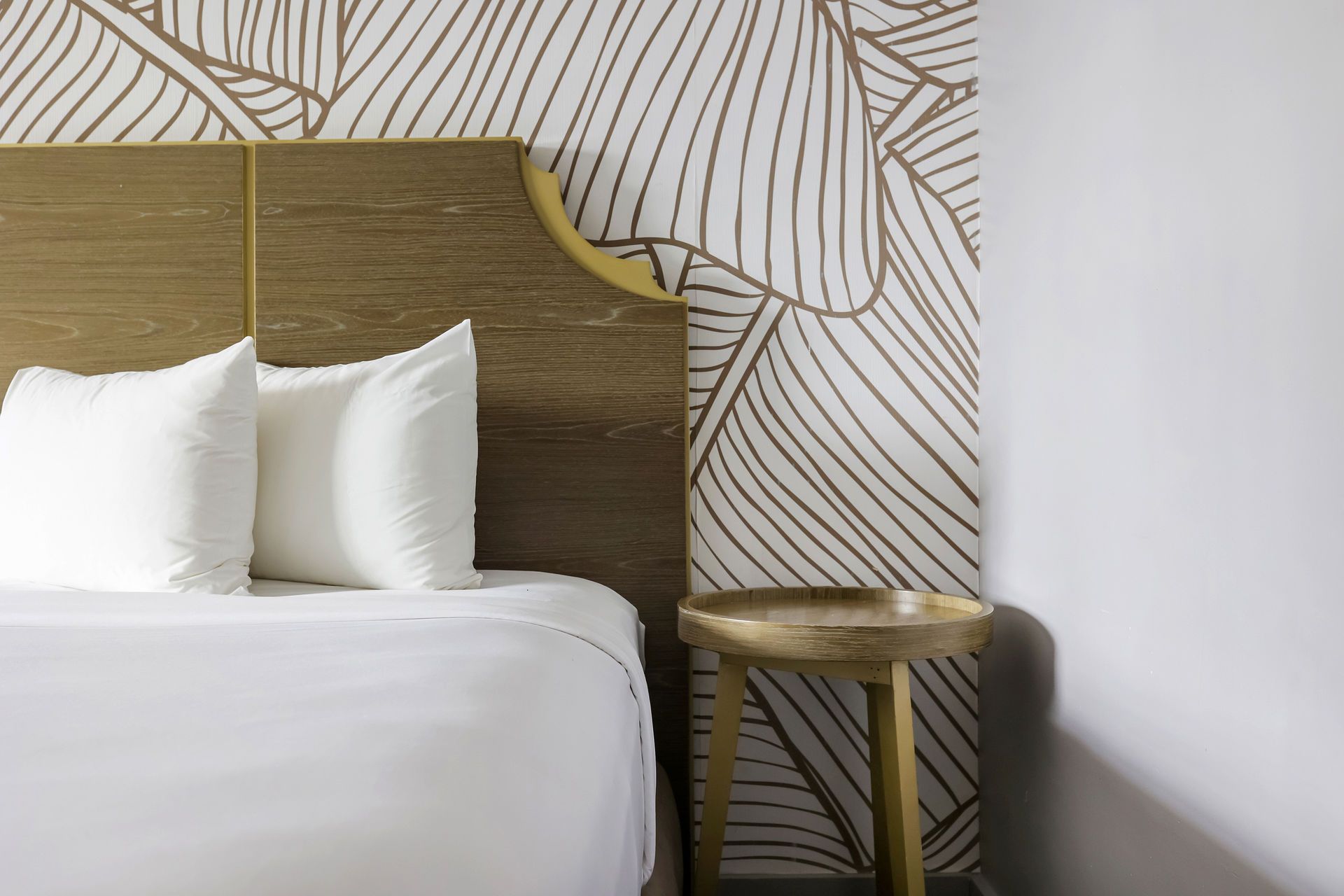 A hotel bed with white pillows and bedding, a light wood headboard, and a wall with brown and white leaf-patterned wallpaper.
