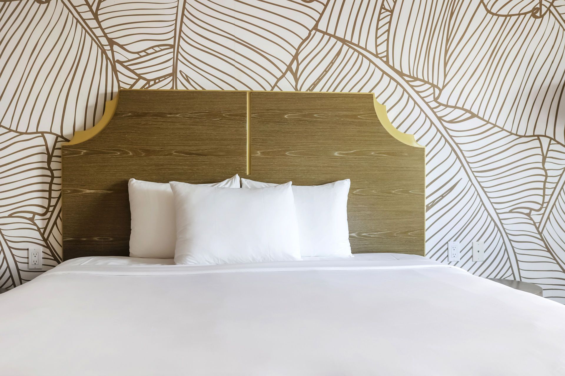 Bed with white linens, three pillows, wooden headboard, and banana leaf wallpaper background.