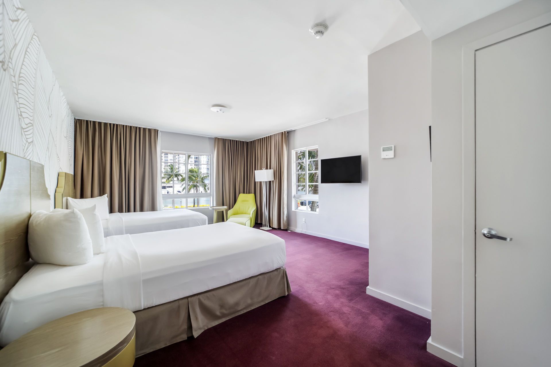 A hotel room with two beds, beige curtains, a lime-green accent chair, a wall-mounted TV, and purple carpeting.
