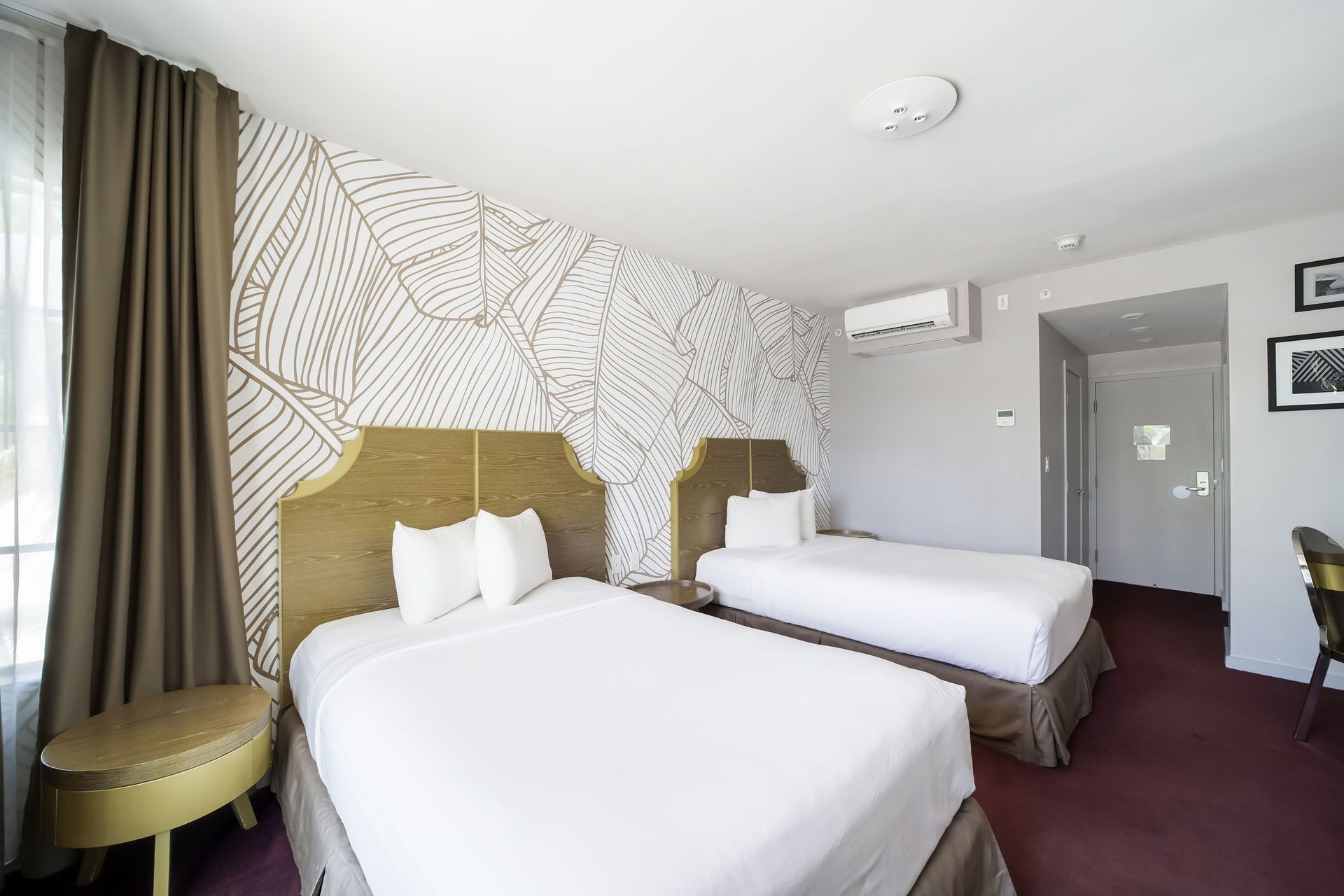 A hotel room with two beds, beige upholstered headboards, leaf-patterned accent wall, brown curtains, and carpet.
