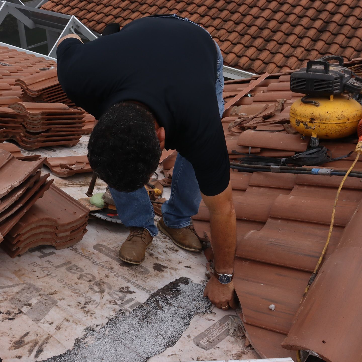 A person in a black shirt and jeans kneels on a residential roof, working with terracotta tiles and roofing equipment.