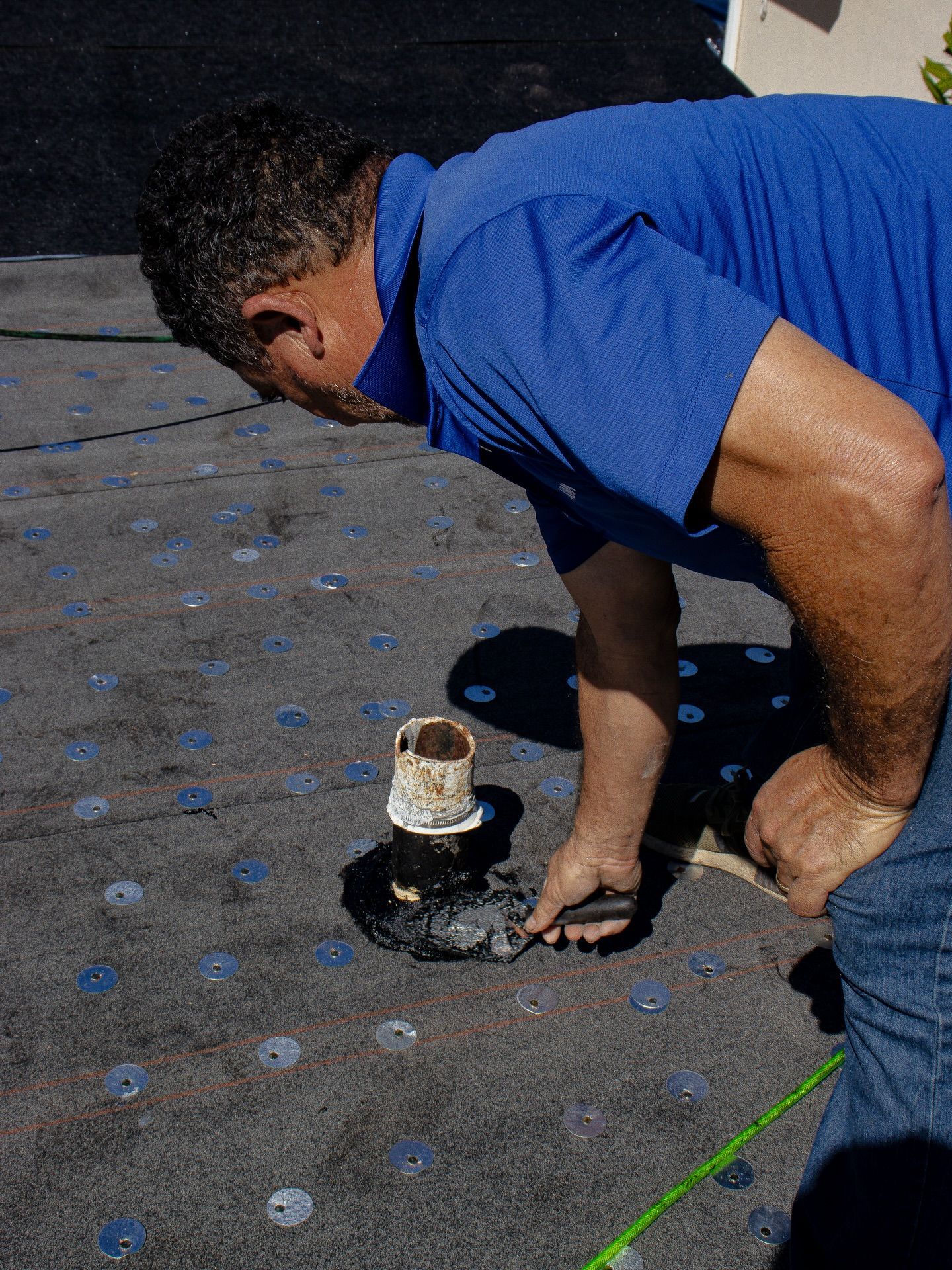 A person in a blue shirt applies sealant around a plumbing vent pipe on a flat, dark roof with many circular fasteners.