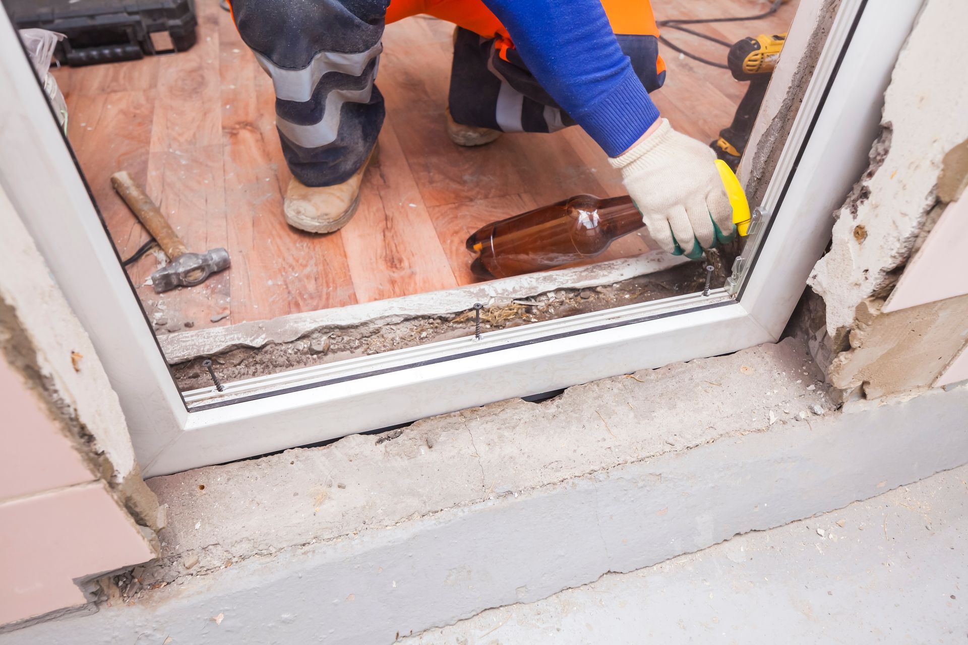 A worker in gloves installs a window frame into a wall opening, with tools nearby on a wooden floor.