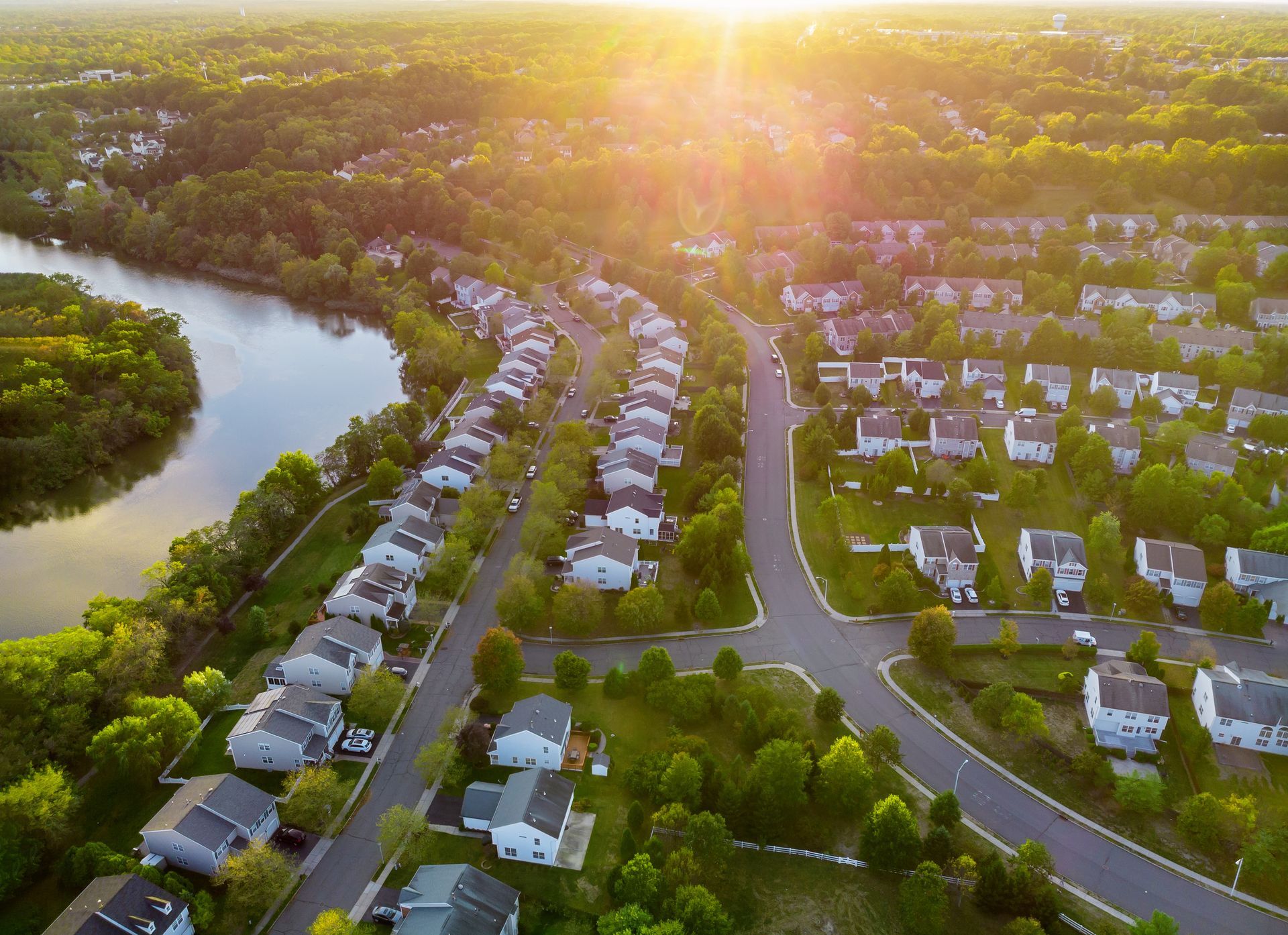 Aerial view of a residential neighborhood with suburban houses nestled among green trees beside a winding river at sunset.